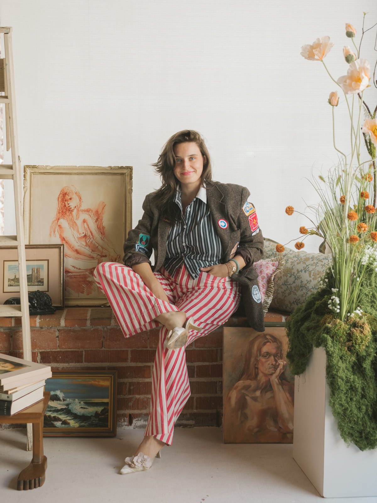 Young woman with wavy brown hair sitting cross-legged on a brick ledge surrounded by framed artwork, including a sketch of a seated woman and a seascape painting, next to a large arrangement of flowers and greenery in a white vase.
