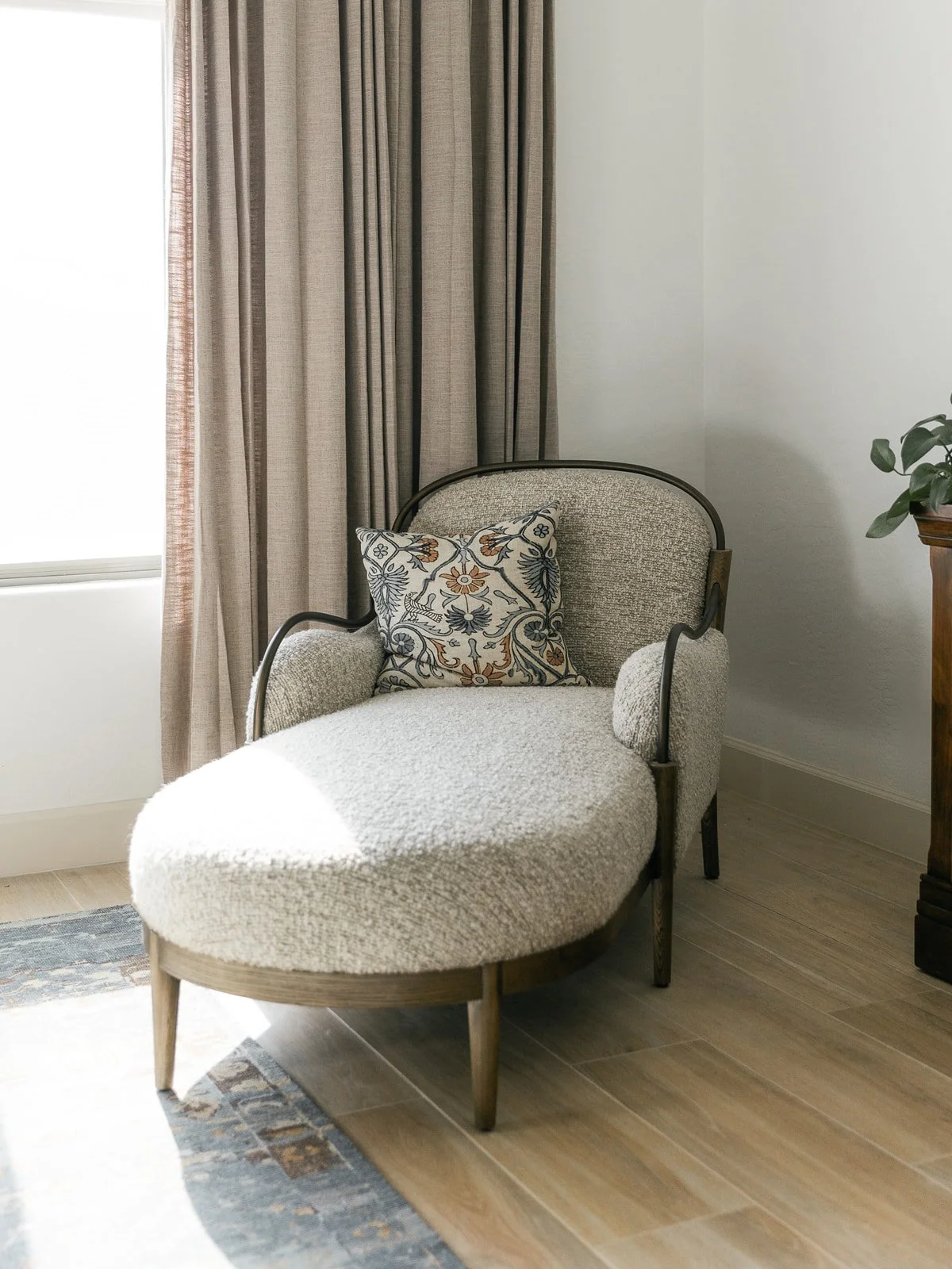 A cushioned armchair with a patterned throw pillow, next to a window with beige curtains and a potted plant on a side table.