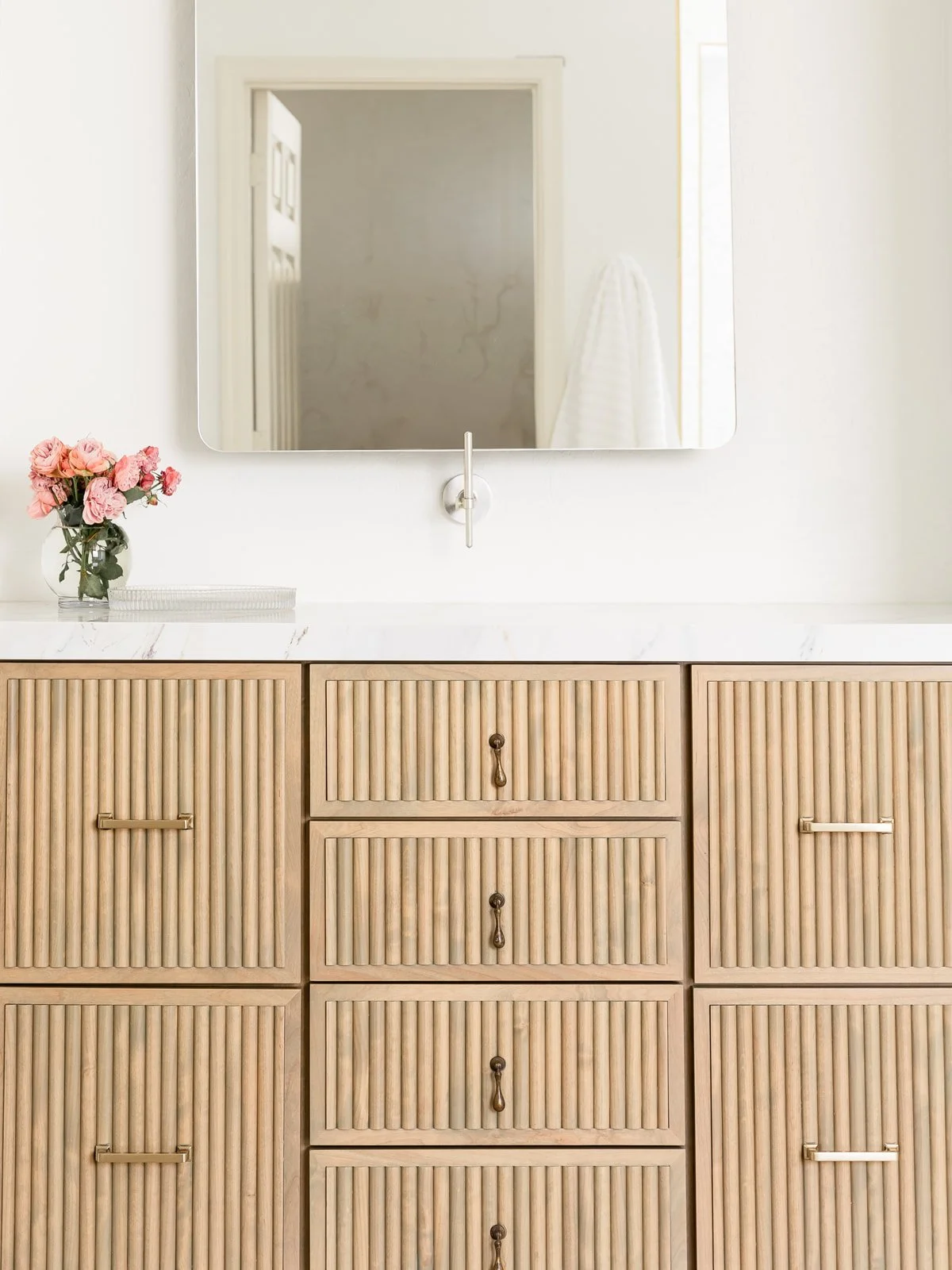 Bathroom vanity with a mirror, a vase of pink flowers, and a white marble countertop. The vanity features wooden cabinet doors and drawers with vertical slats and small handles. A towel hangs on the wall in the reflection of the mirror.