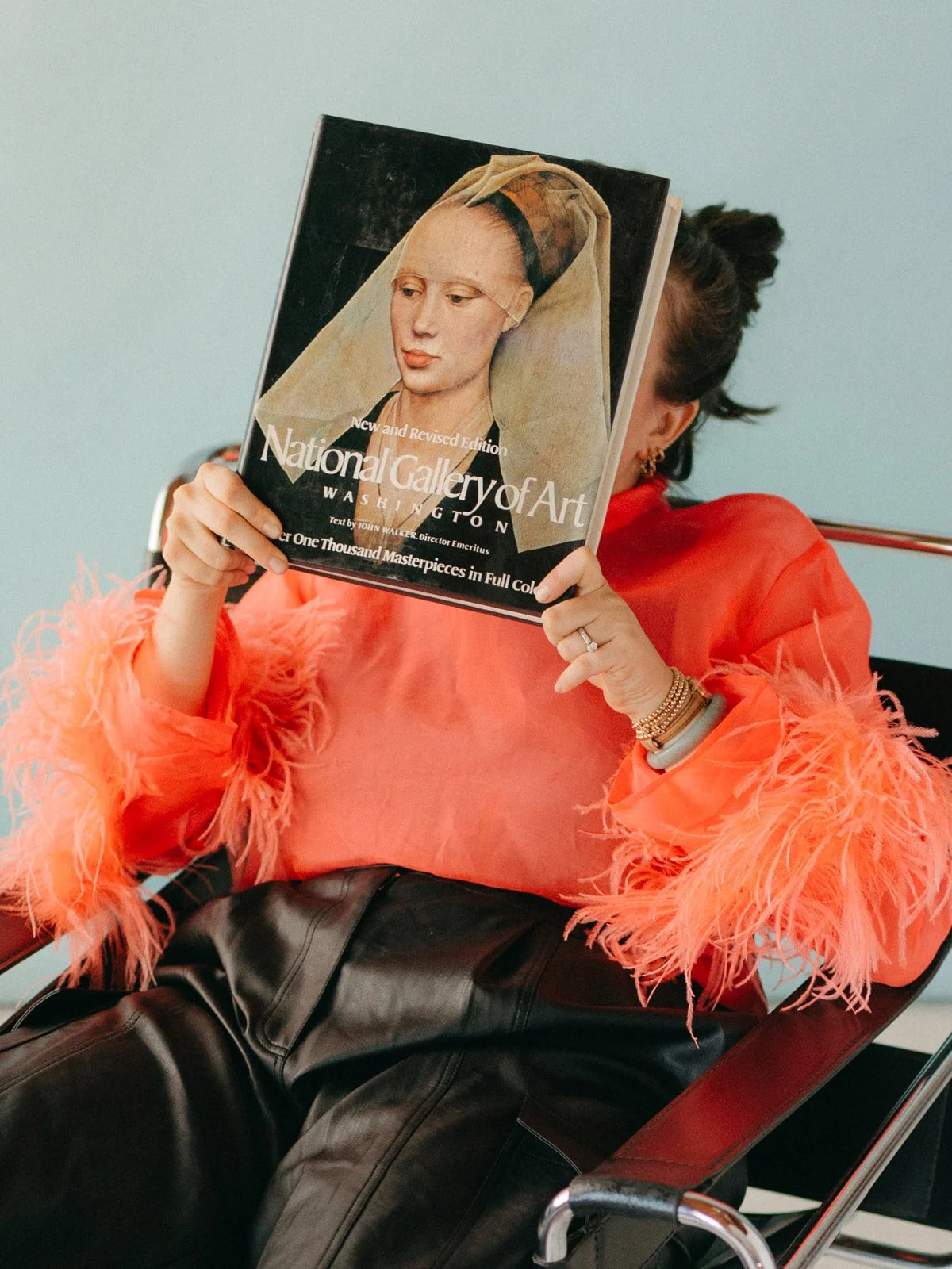 A woman with dark hair tied back, wearing a coral-colored top with feathered sleeves and black leather pants, sitting in a chair and holding a copy of the National Gallery of Art magazine in front of her face.