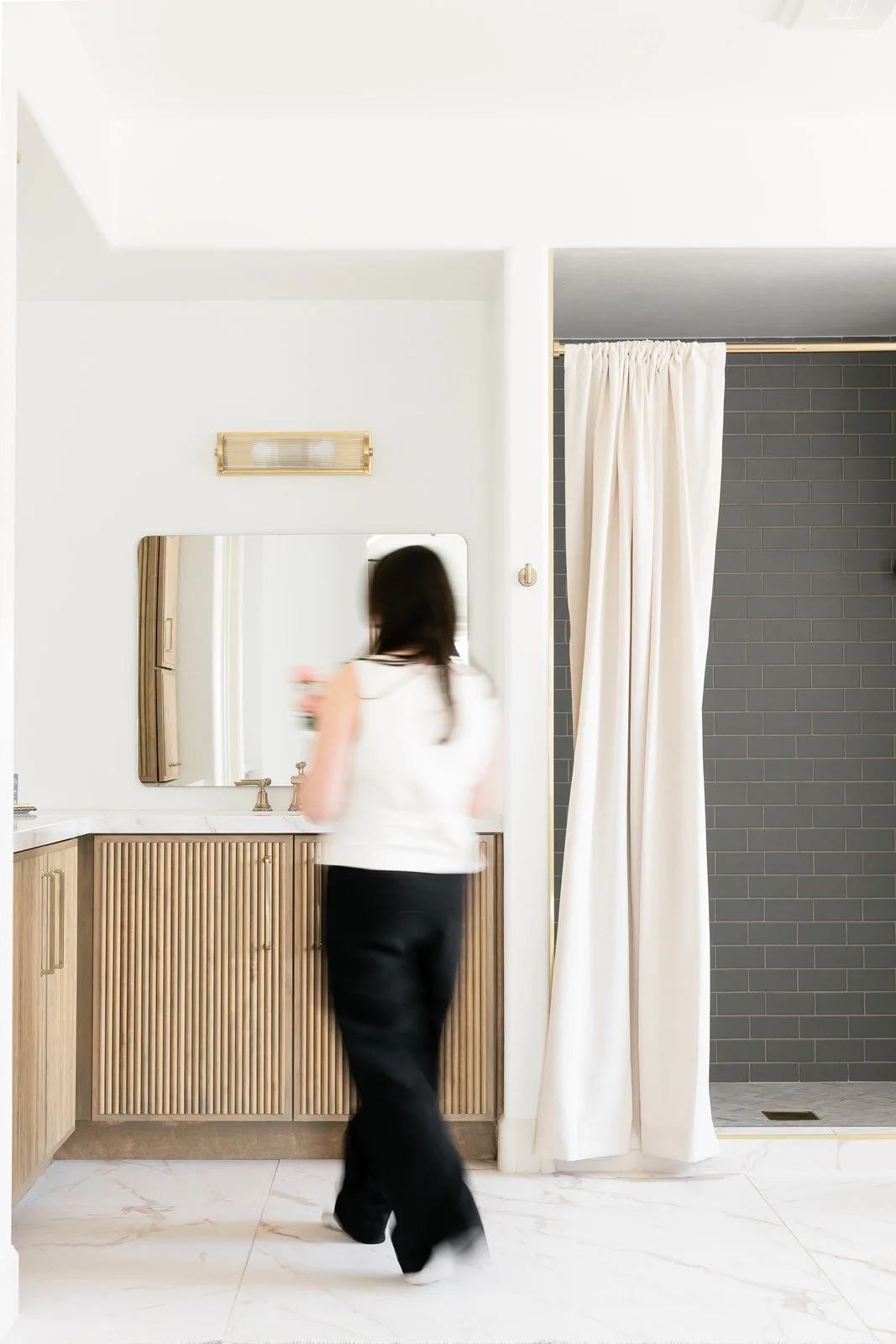 A woman with long dark hair, wearing a white top and black pants, standing at a bathroom vanity with a mirror, holding a pink object, with a shower curtain to her right and a gray tiled shower area behind it.