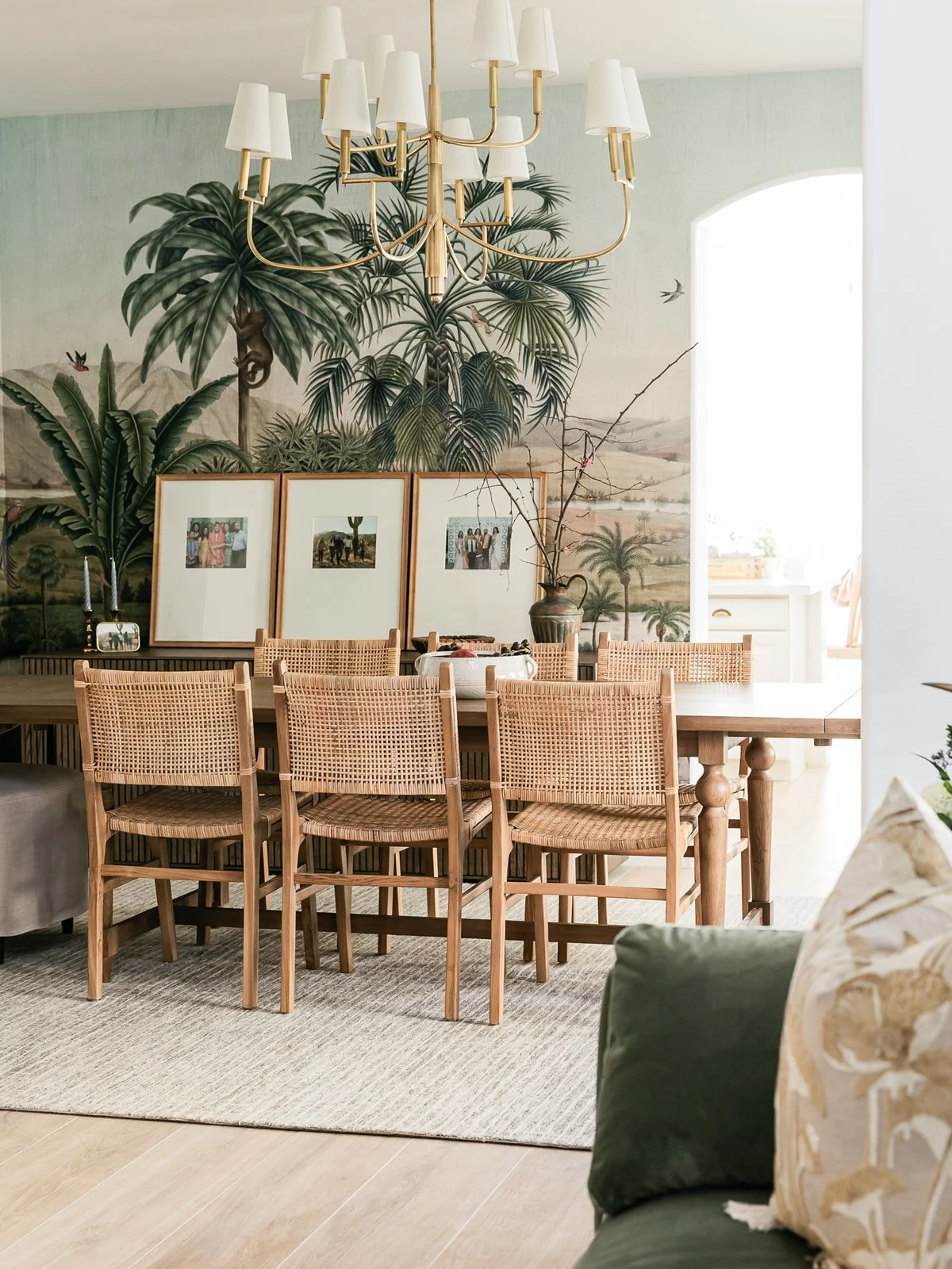 A dining room with a large wooden table surrounded by six woven rattan chairs. The wall behind the table features a mural of lush tropical plants and trees. There are three framed photographs leaning against the wall behind the table. A chandelier wi