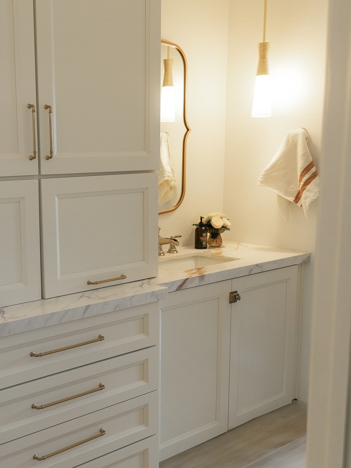 A bathroom vanity with white cabinets and drawers, a marble countertop, a small sink, a mirror, and floral decor, illuminated by two hanging pendant lights.