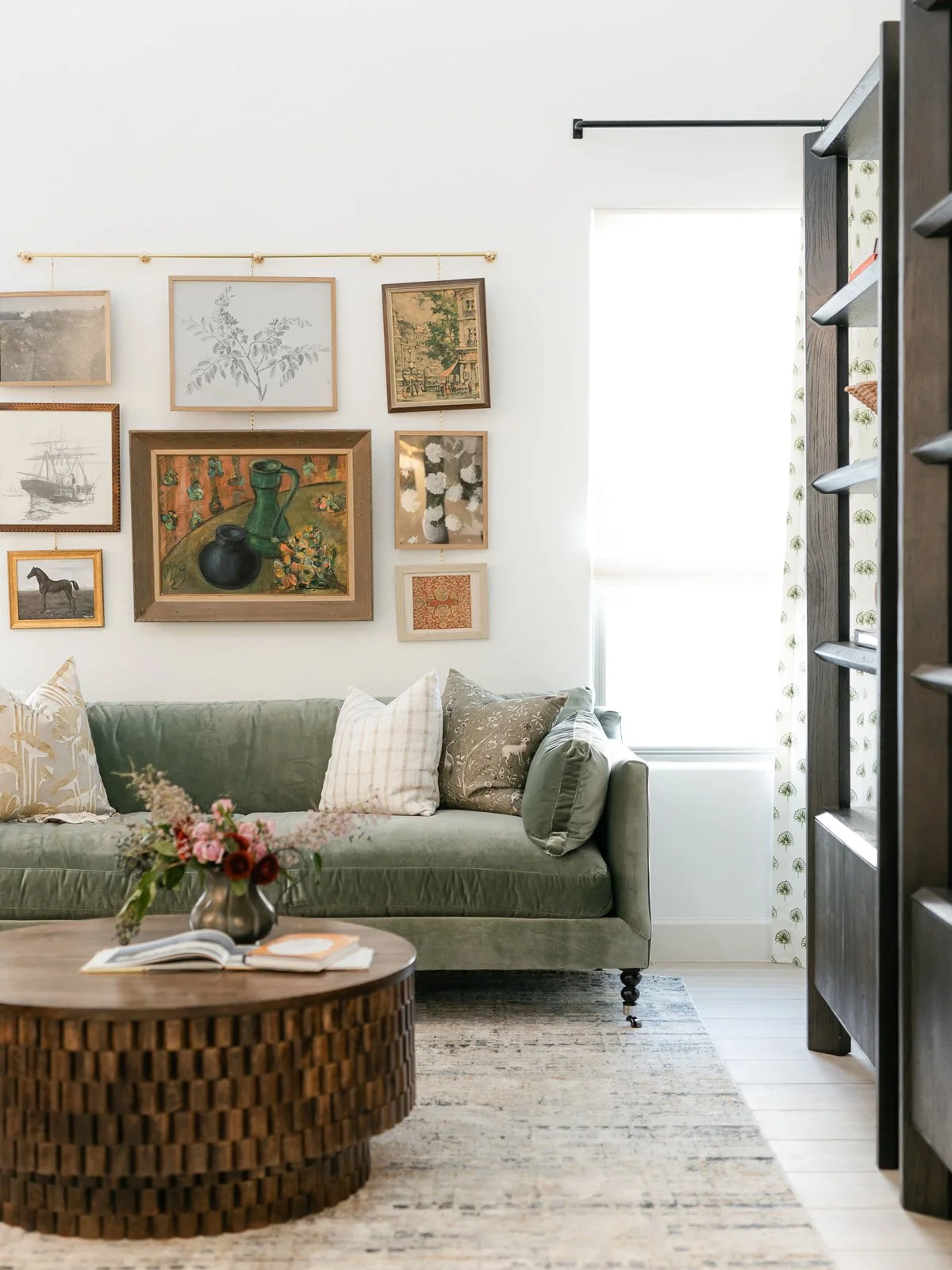 A cozy living room with a green sofa adorned with several pillows, a round wooden coffee table with a vase of flowers and an open book, a colorful gallery wall of framed artwork behind the sofa, a large window with patterned curtains, and a light-col