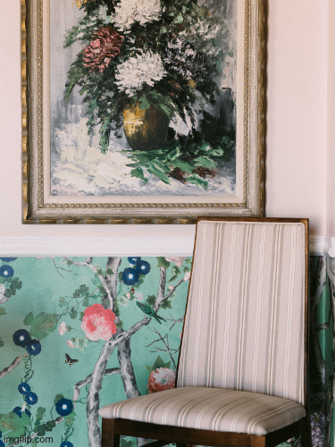 A framed floral painting hangs above a striped upholstered chair in a room with a decorative wall and a painting of a tree and flowers.