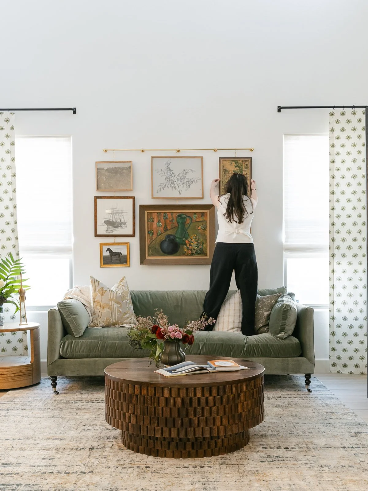 A woman standing on a green sofa adjusting pictures on the wall in a bright living room with white walls, windows with white blinds, a wooden coffee table with a vase of flowers, and various wall art.