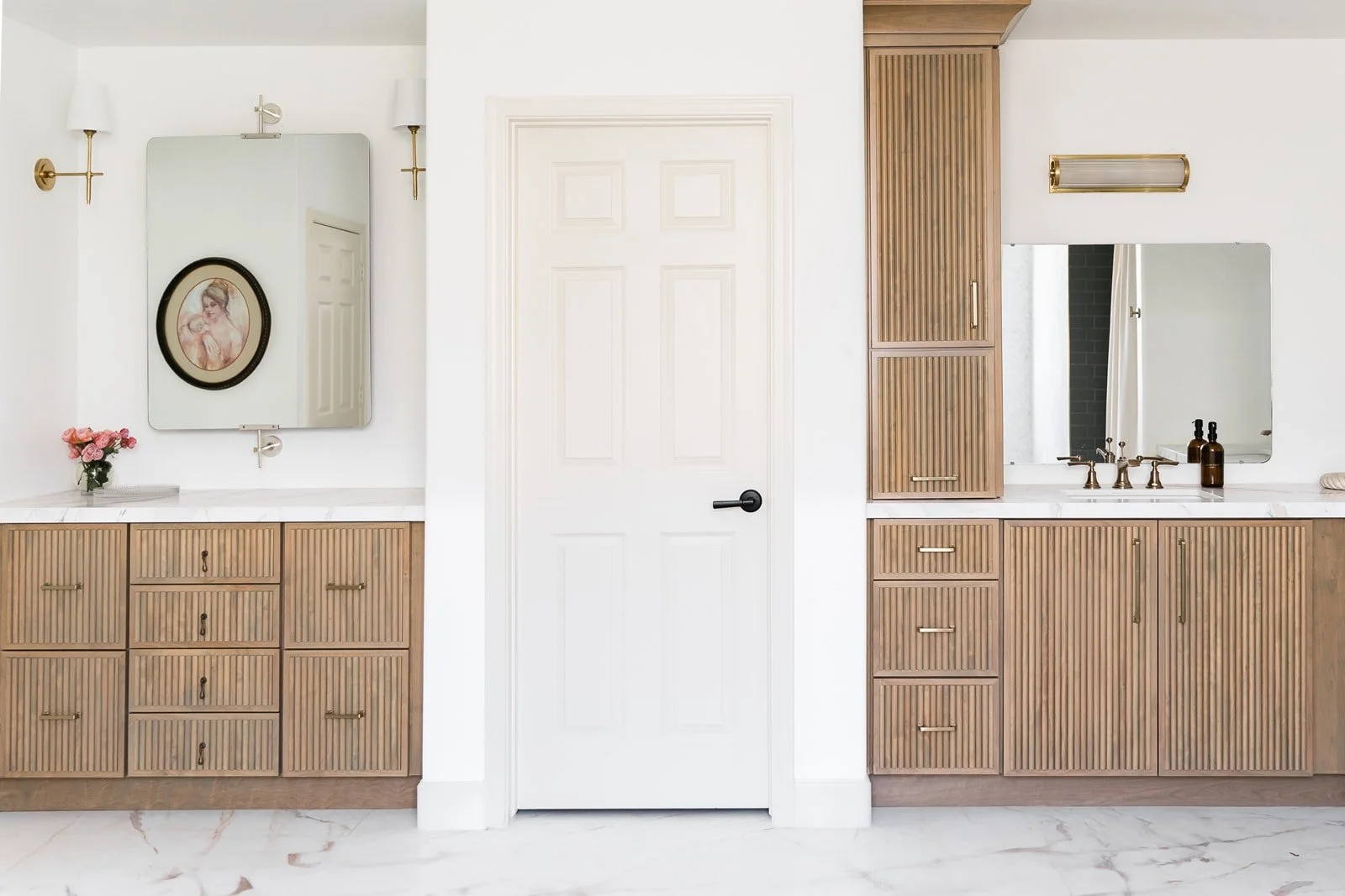 A bathroom with two wooden cabinetry vanities, each topped with a white marble counter and a mirror. The left vanity has a small flower arrangement and pink flowers, while the right has soap dispensers. A white door with a black handle is centered be