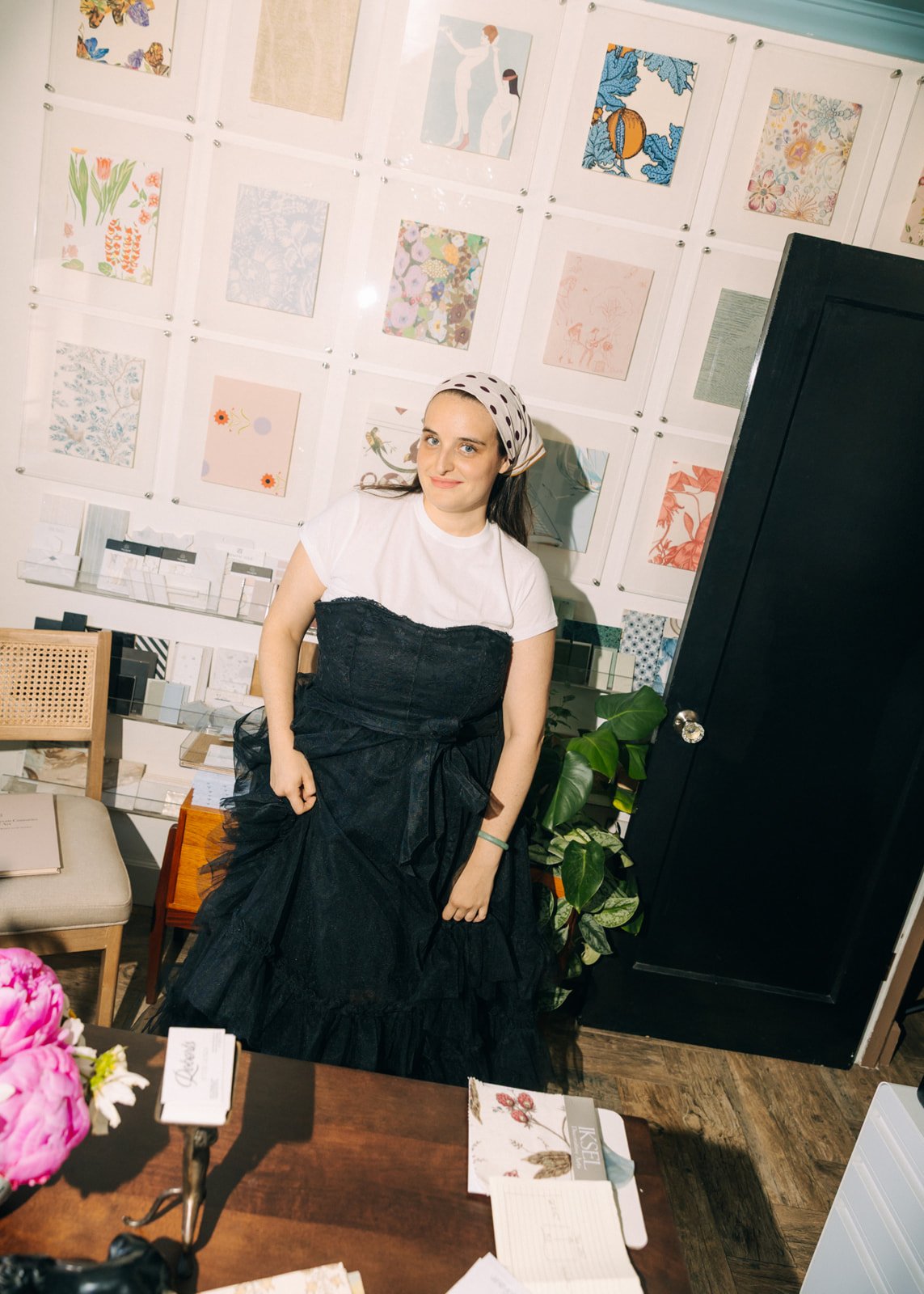 A woman stands in a decorated room with colorful posters or artwork on the wall behind her. She is wearing a white t-shirt, a black dress or apron, and a polka dot headscarf. There are chairs, plants, and various papers or swatches on the table in front of her.
