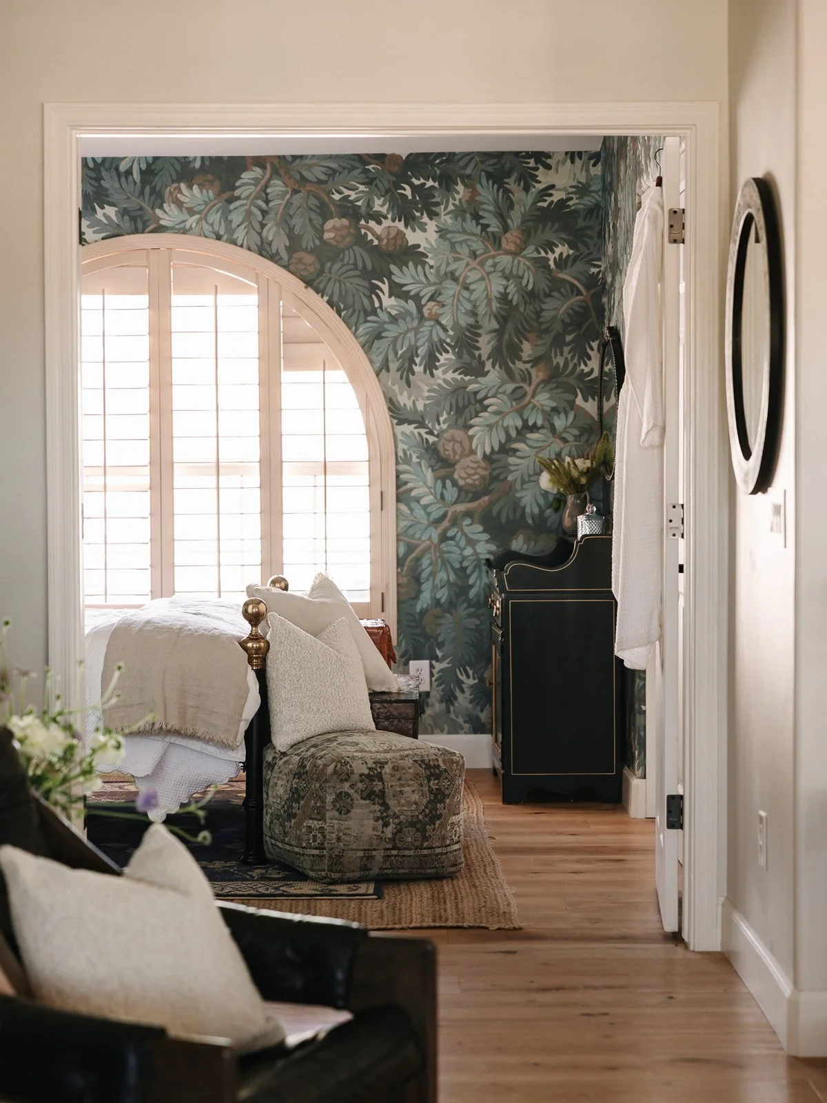 Interior view of a bedroom with wall decor featuring leafy patterns, a window with wooden shutters, vintage furniture, and a hardwood floor.