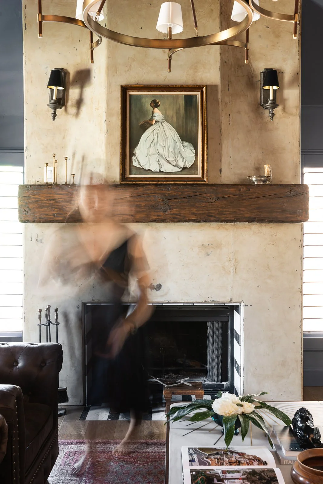 Blurred person moving inside a living room with a fireplace, artwork of a woman in a wedding dress above the mantel, and a modern chandelier hanging from the ceiling.