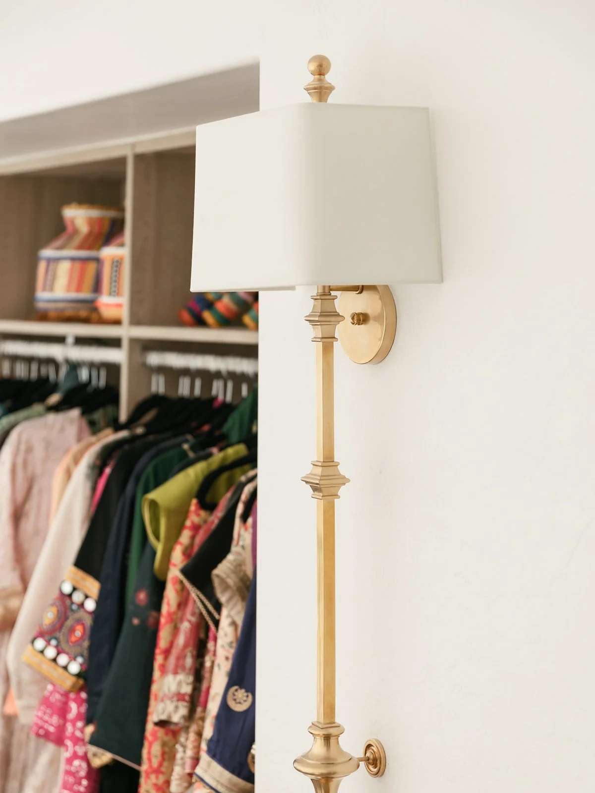 Wall-mounted gold-colored sconce with a white shade, next to a closet with clothes and shelves of books.