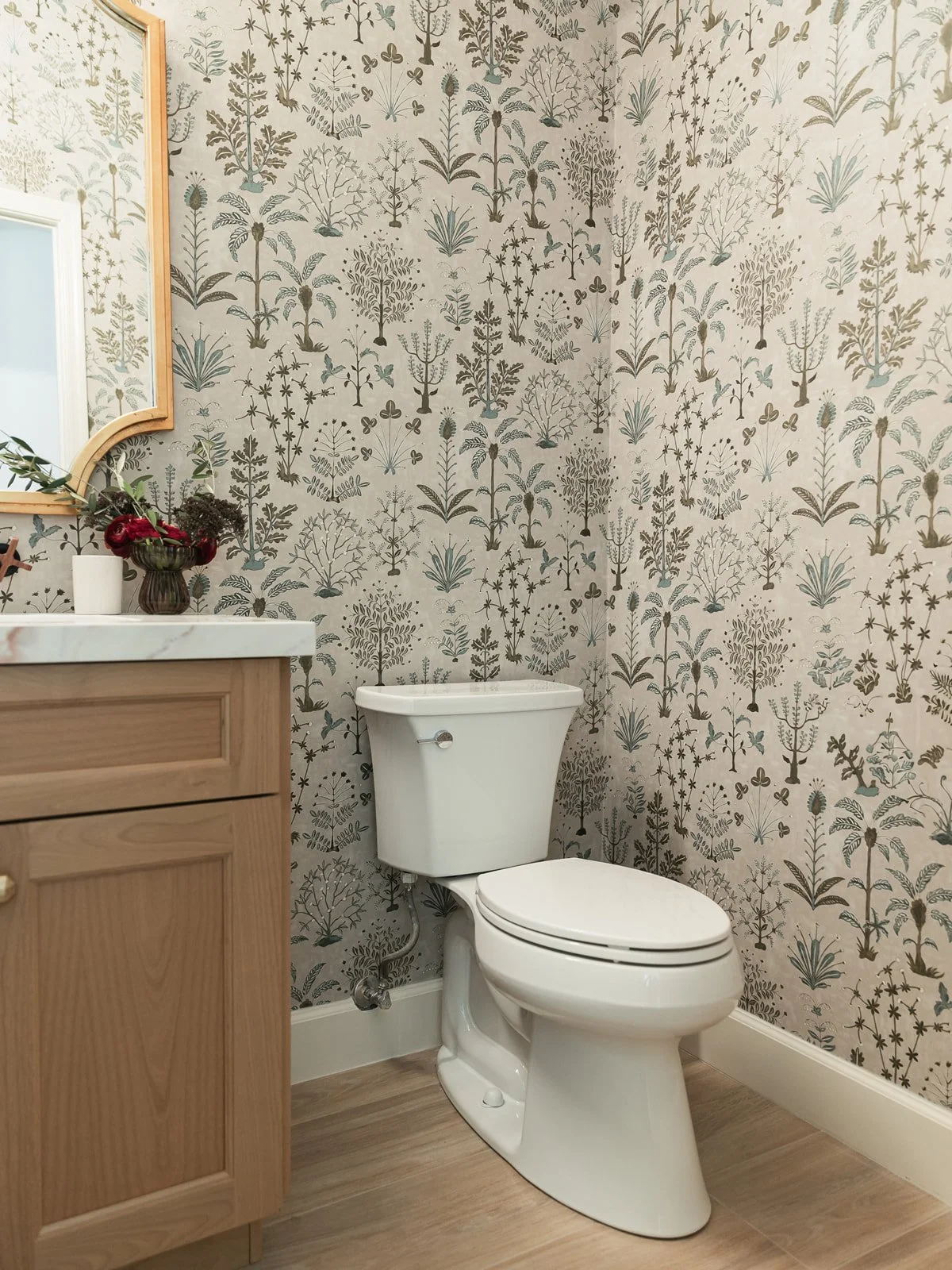 A bathroom with beige and gray floral and plant patterned wallpaper, a white toilet, a wooden vanity with a white countertop, and a mirror with a wooden frame. There are small decorative items and a vase with flowers on the vanity.