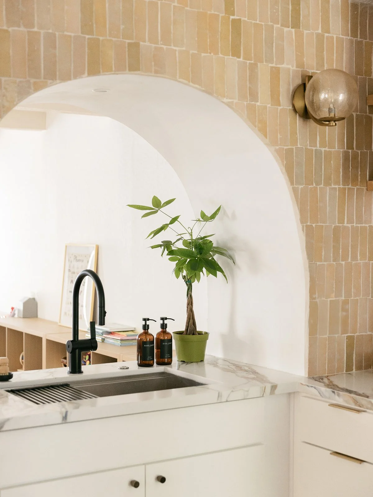 A kitchen with white cabinets, a marble countertop, a black faucet, a potted plant, and two brown soap bottles. There's an arched opening with beige brick wall and a round wall sconce.