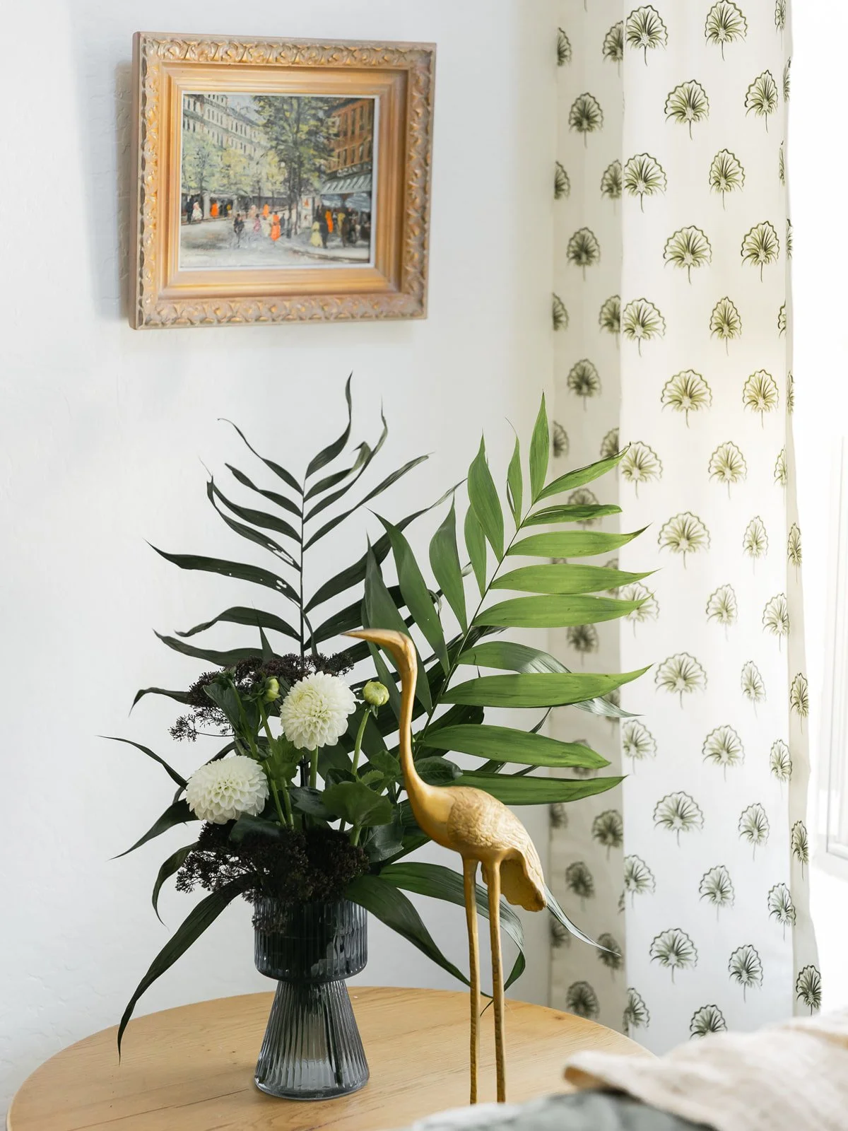 A floral arrangement with green leaves, white flowers, and a gold-colored heron sculpture in a dark glass vase on a wooden table, with a framed painting of a city street scene and a white curtain with green ginkgo leaf patterns in the background.