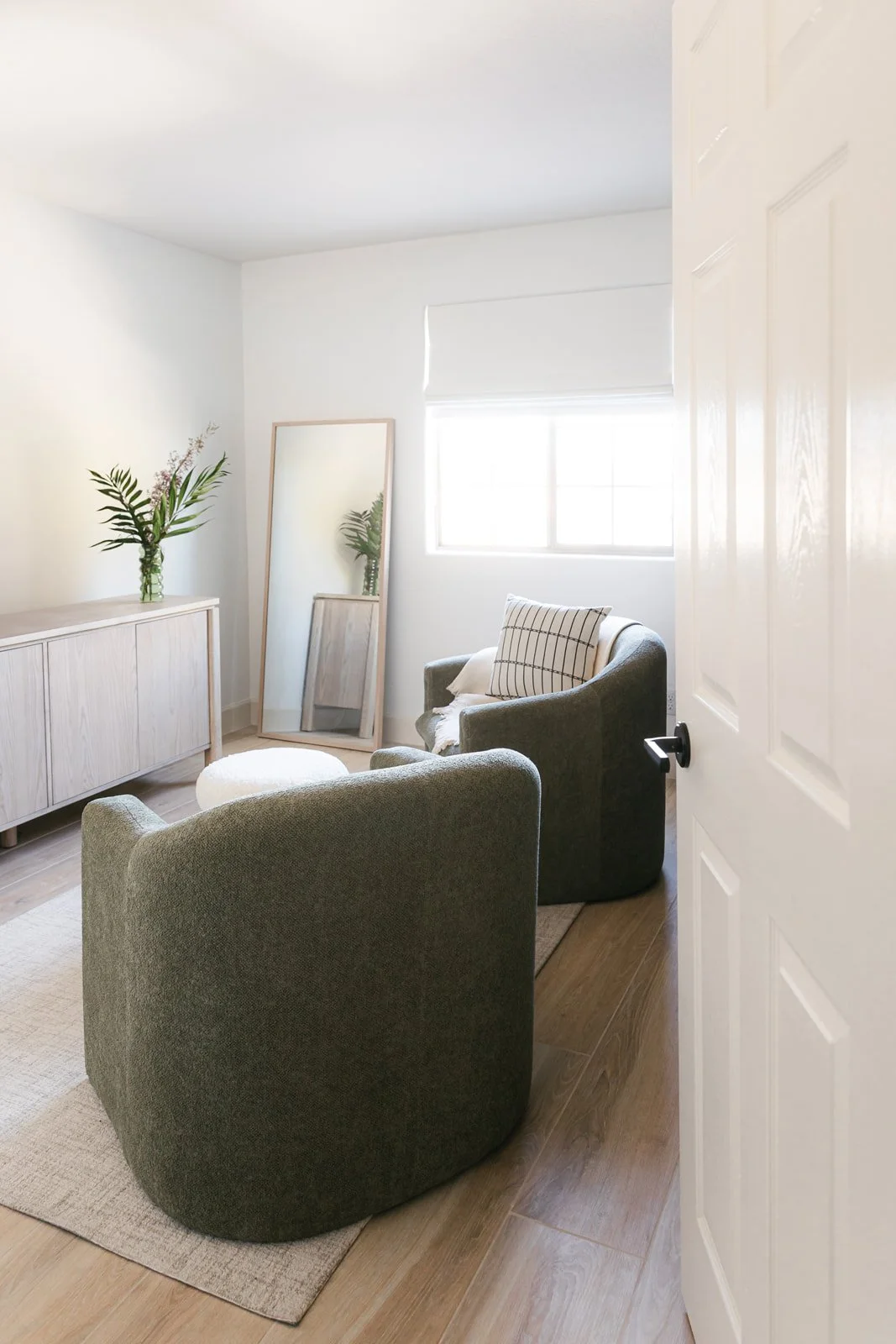 A cozy, minimalistic living room with two dark green armchairs, a round ottoman, a wooden sideboard with a vase of flowers, a large mirror leaned against the wall, and a window with a white shade, all with light-colored walls and wooden flooring.