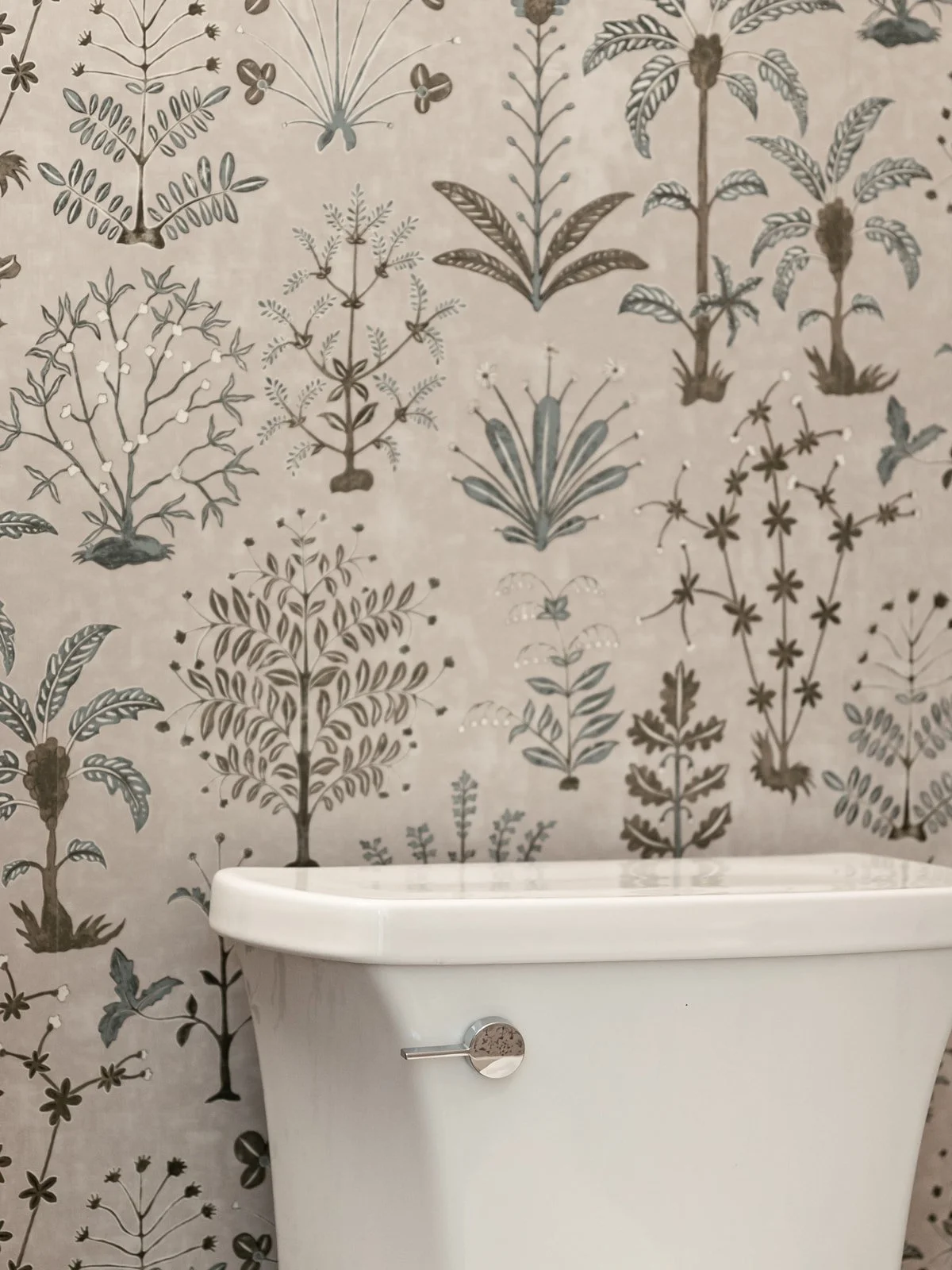 Close-up of a white toilet tank with a chrome flush handle, set against a wallpaper with botanical illustrations of various stylized plants and flowers.