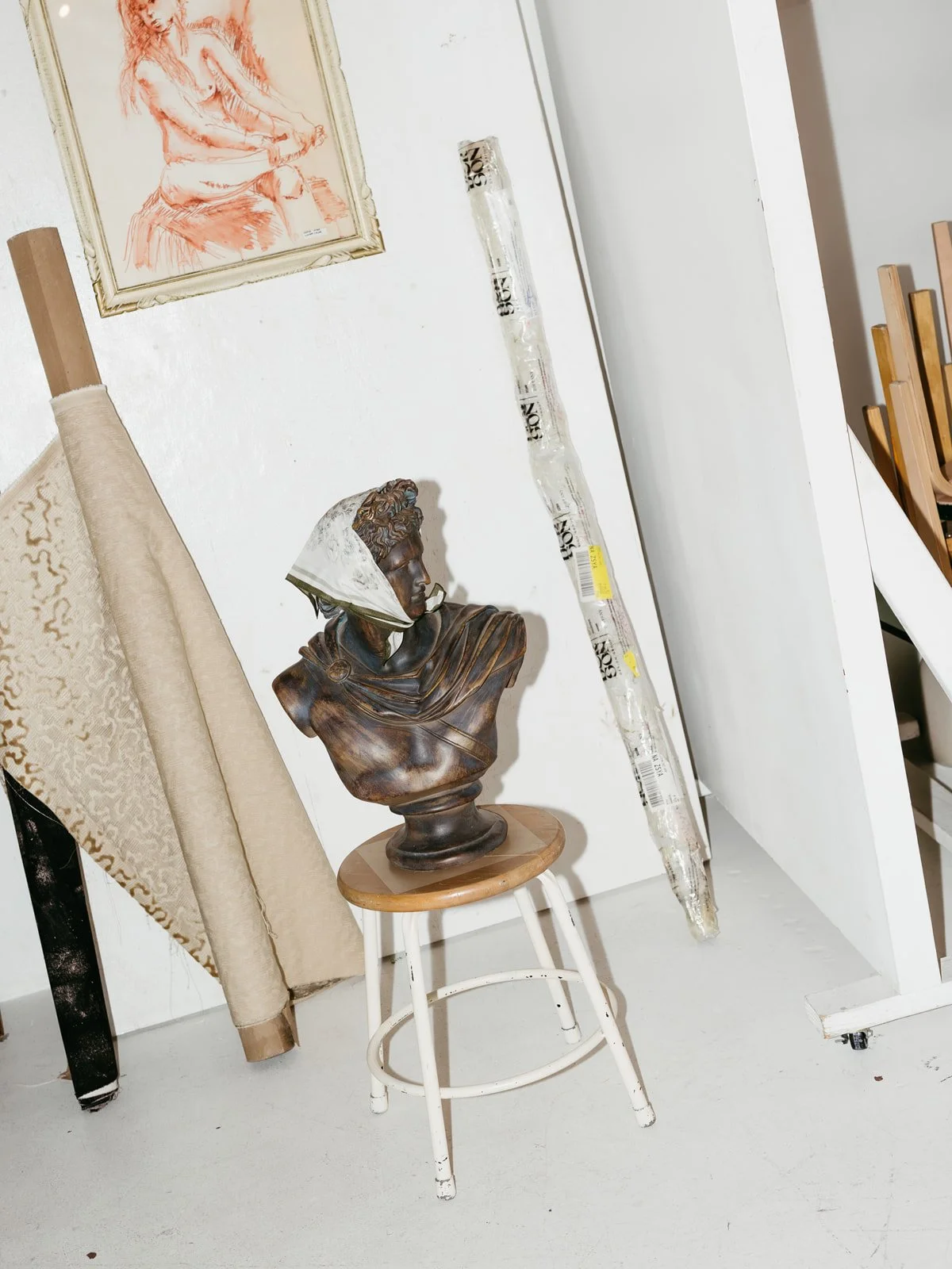 Art studio with a bust sculpture on a stool, a rolled-up fabric or wallpaper, a framed sketch of a seated woman, and a long object wrapped in plastic leaning against the wall.