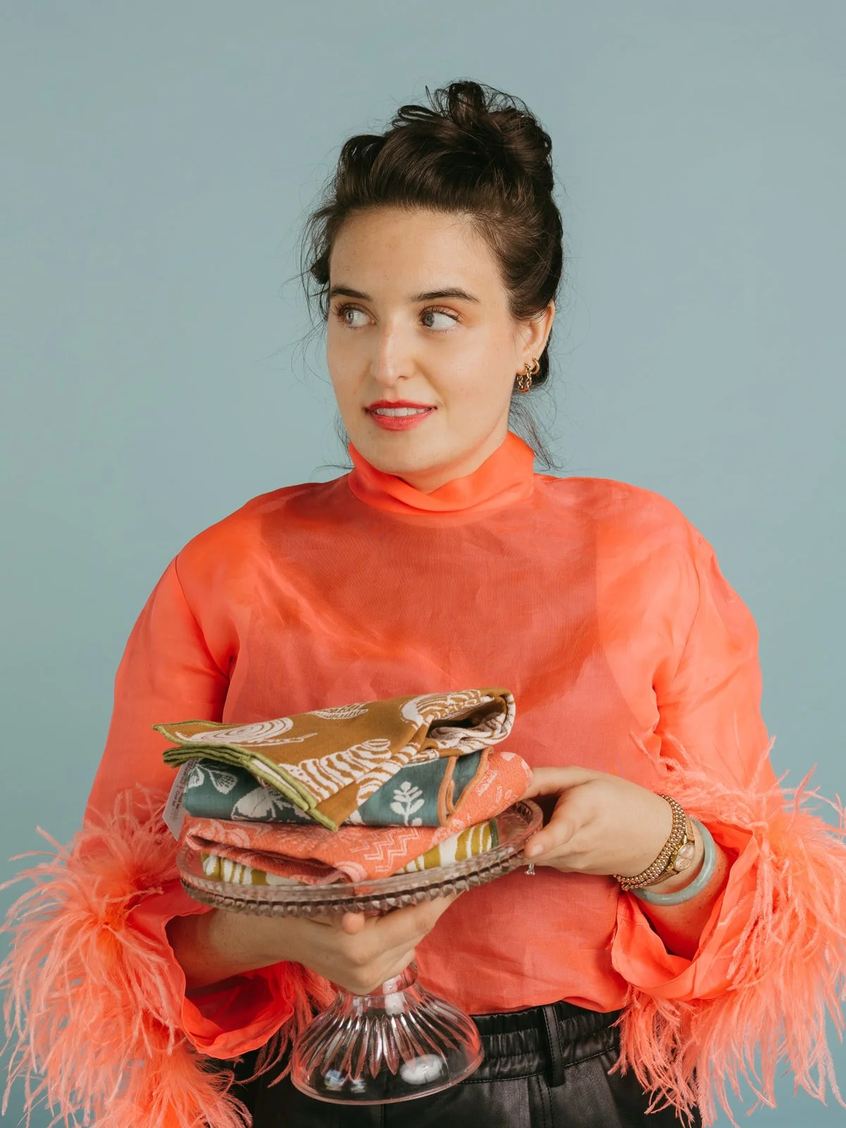 A woman in an orange, sheer top with feathered sleeves holding a glass cake stand with folded colorful cloth napkins or fabric pieces against a pastel blue background.