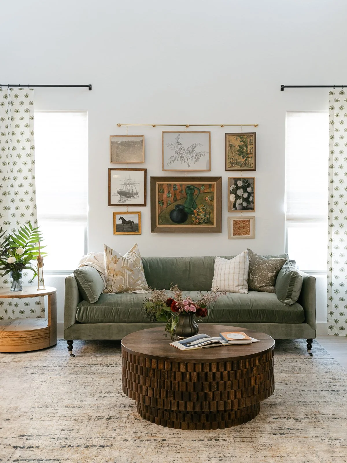 A living room with a green sofa, decorative pillows, a round wooden coffee table with a vase of flowers, and wall art, lights, and curtains in the background.