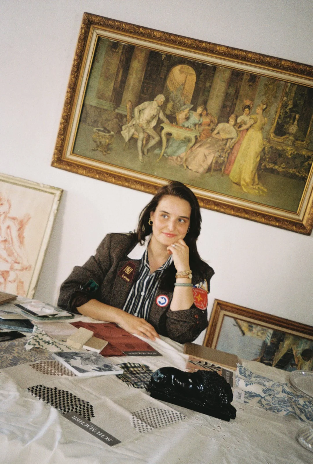 A woman with dark hair, wearing a brown blazer with patches, sitting at a table with various fabric samples, papers, and a black item. She is resting her chin on her left hand and looking slightly to her right. Behind her are framed paintings, including a large classical painting on the wall depicting a group of people in an elegant setting.