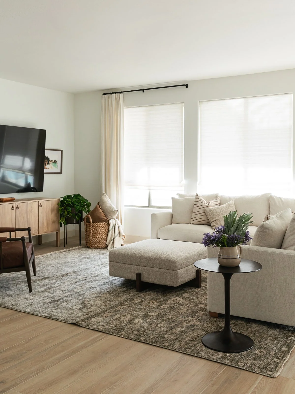 Living room with a beige sofa, matching ottoman, a black side table with a vase of purple flowers, a large TV on a wooden cabinet, and windows with sheer white curtains.