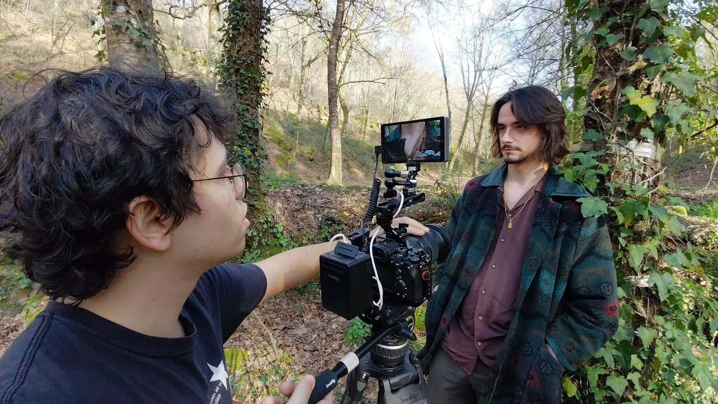 Duas pessoas filmando na floresta com uma câmera de vídeo em um tripé. Uma pessoa ajusta a câmera enquanto a outra observa, vestindo roupas casuais.