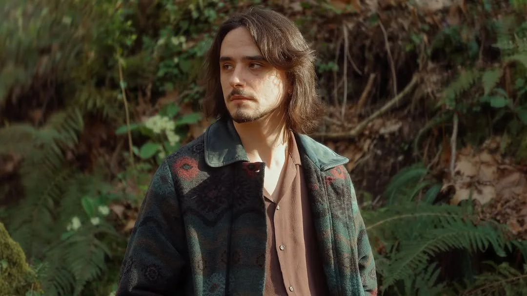 Homem de cabelo castanho claro, com barba por fazer, usando uma jaqueta escura com padrão de xadrez e uma blusa marrom, de pé na floresta