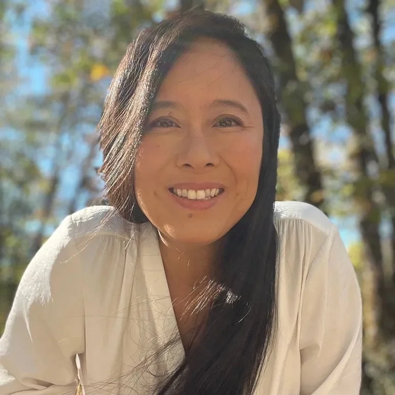 Smiling woman with long black hair outdoors on a sunny day, trees with blurred leaves in the background.