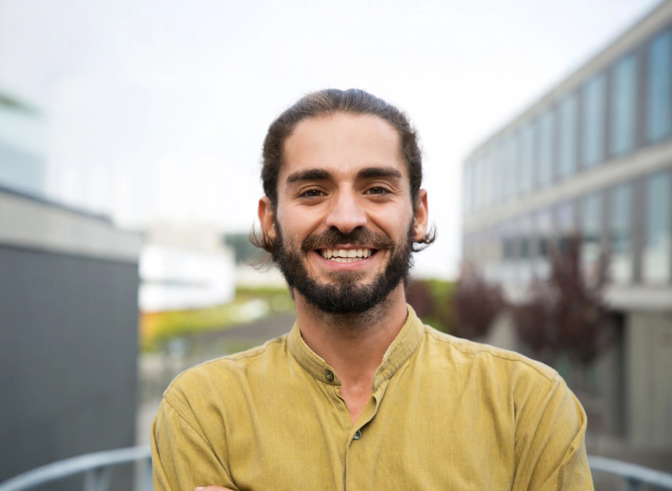 A smiling man with a beard and long hair standing outdoors, wearing a yellow shirt with a blurred urban background.