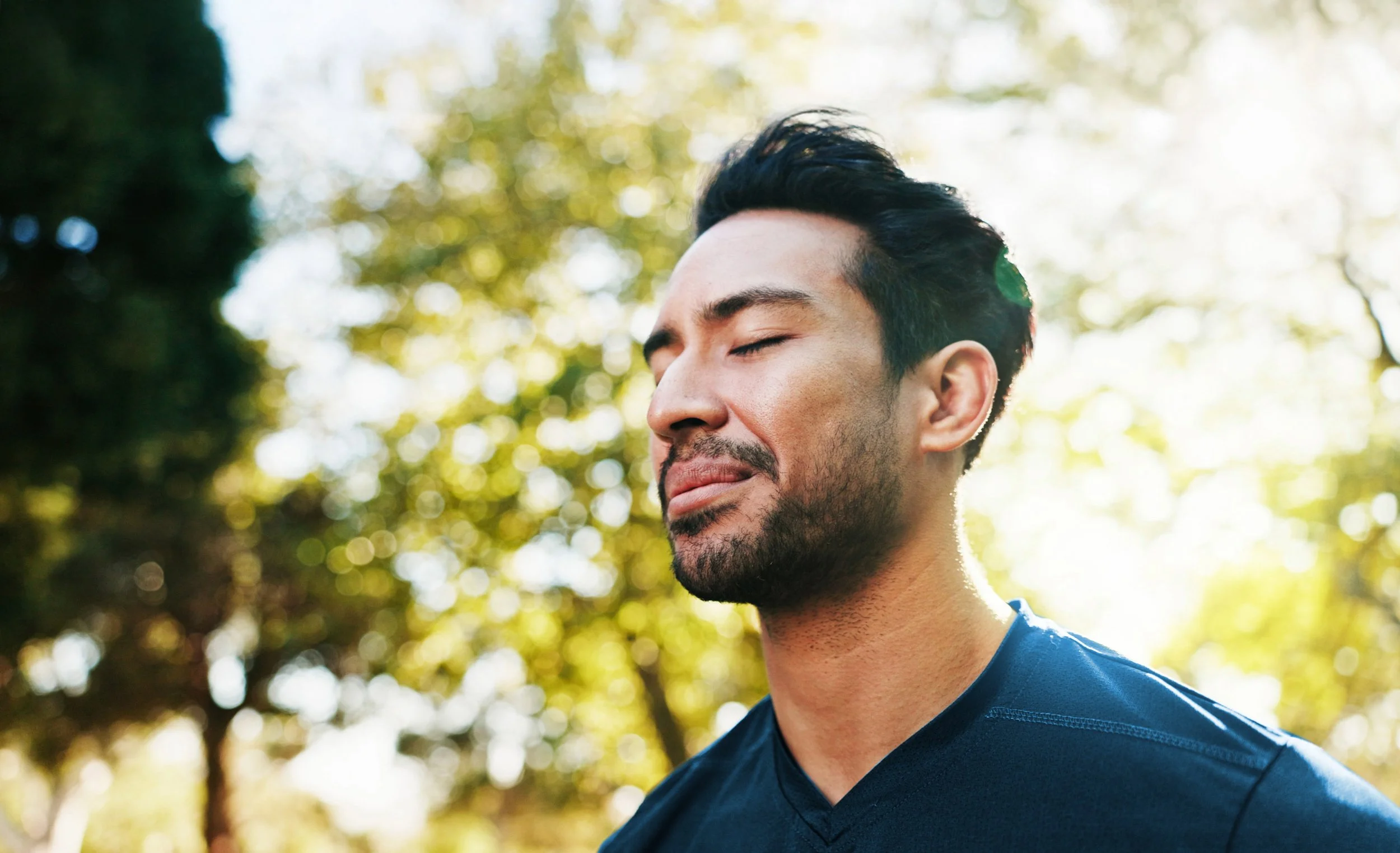 A man with closed eyes and a slight smile outdoors in front of trees with sunlight filtering through.
