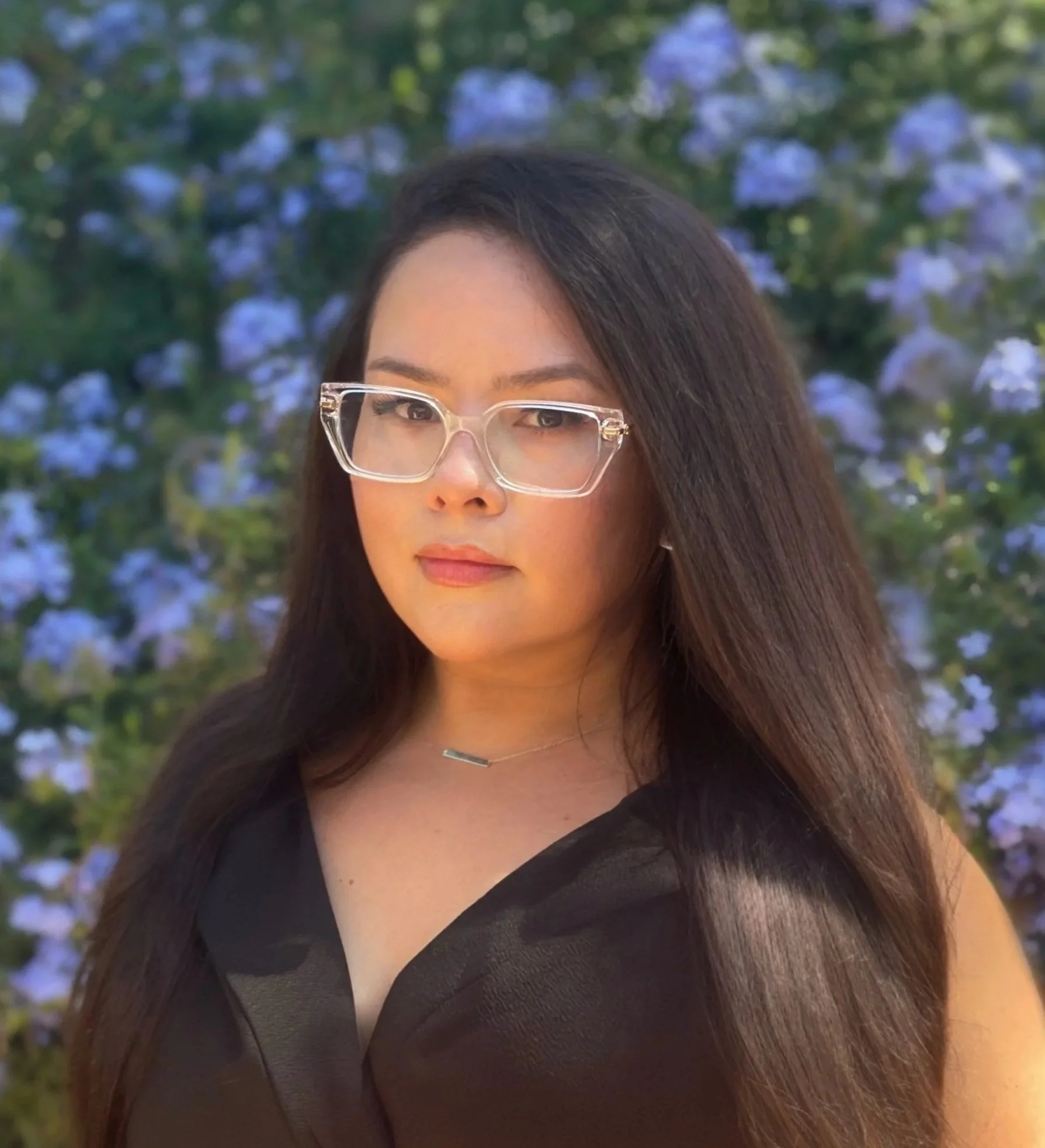 A woman with long dark hair wearing clear glasses and a black sleeveless top, standing outdoors with purple flowers and greenery in the background.