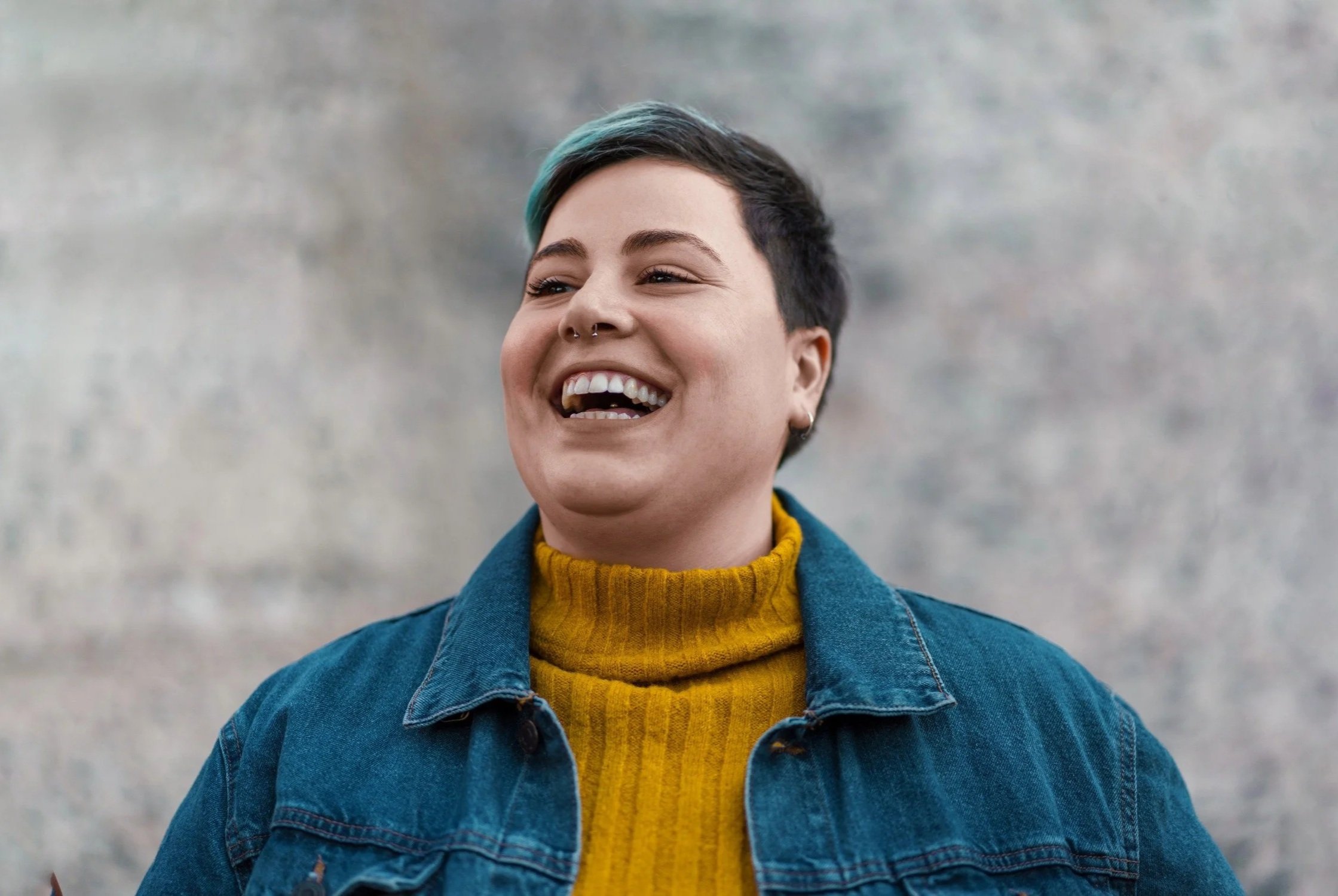 A person with short black hair and blue highlights is smiling, wearing a yellow turtleneck sweater and a blue denim jacket.