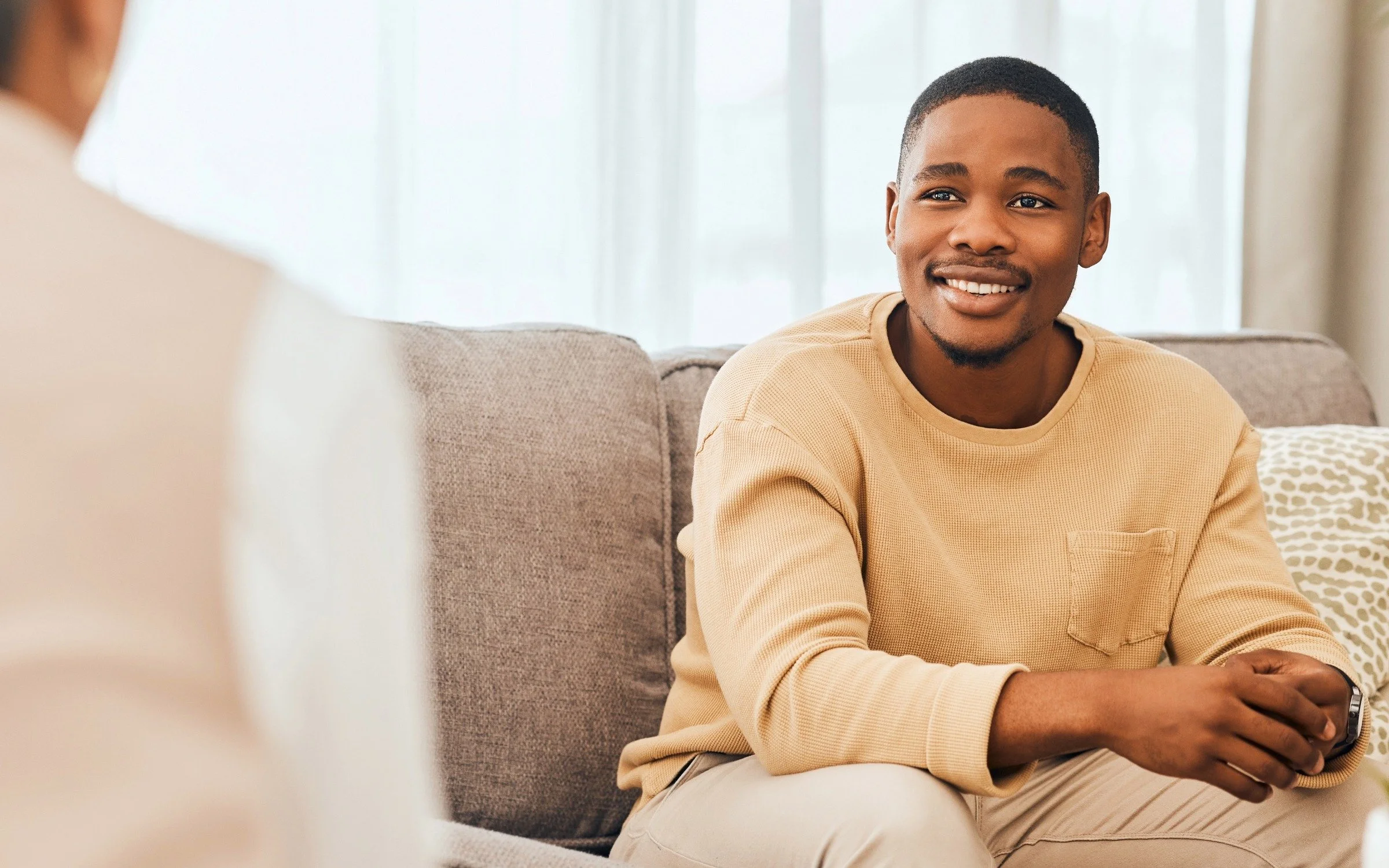 A young man with a smile sitting on a couch in a well-lit room, wearing a beige sweatshirt.