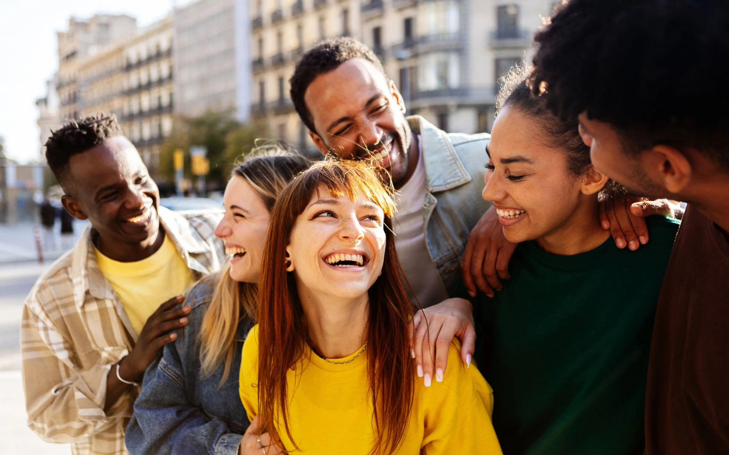 Group of six diverse friends smiling and laughing together outdoors in a city setting.