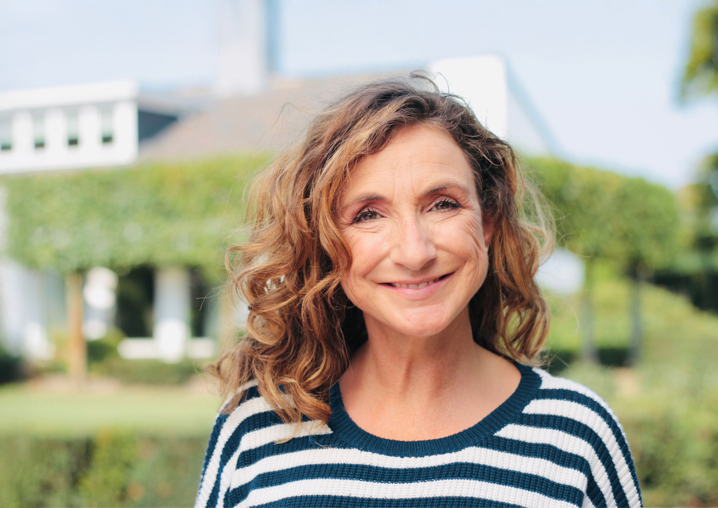 Close-up of a middle-aged woman with curly hair smiling outdoors in a garden, wearing a striped navy and white sweater.