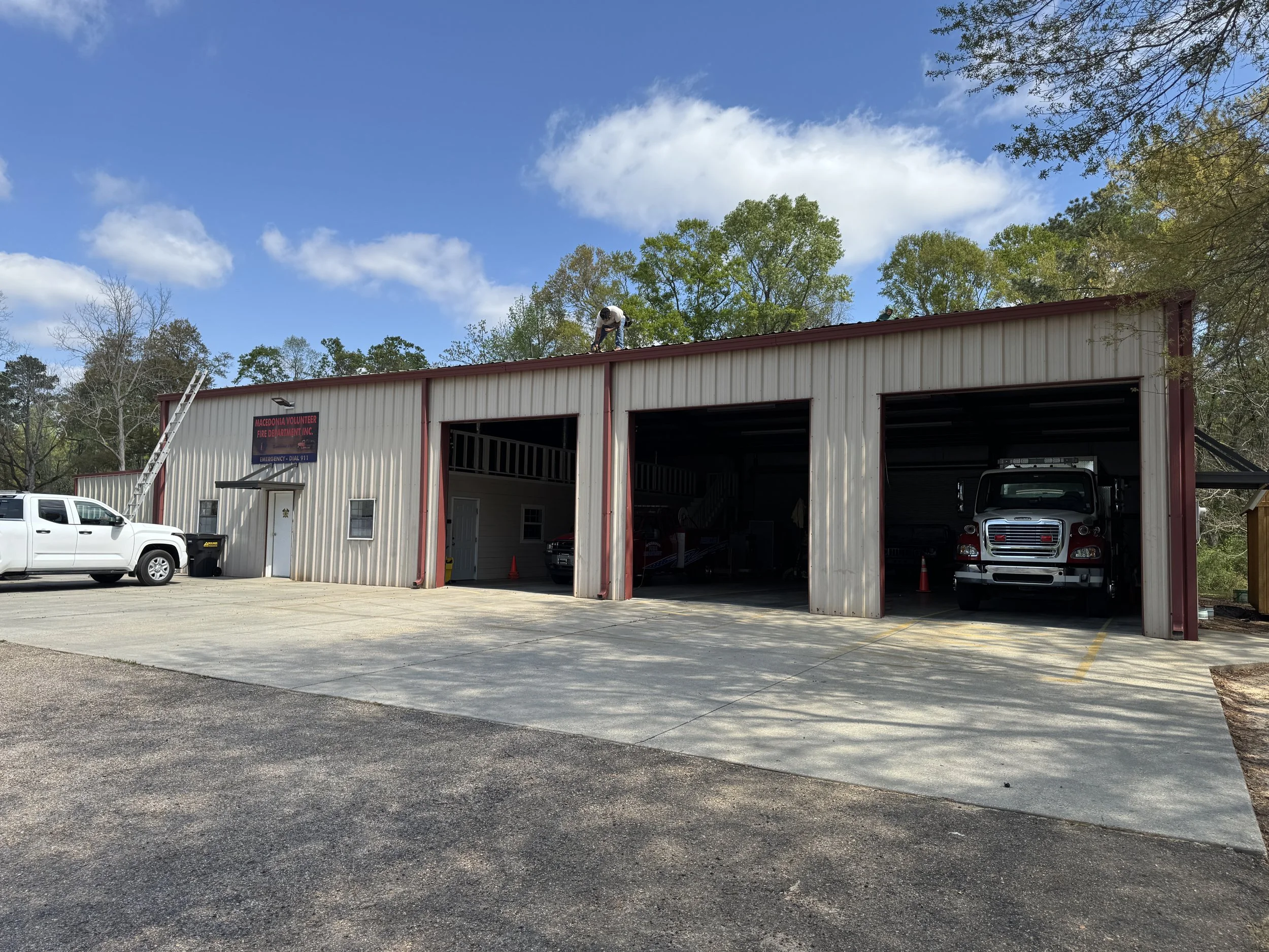 A daytime scene outside of a fire station with three garage bays, a white pickup truck parked outside, and a worker on the roof of the building.