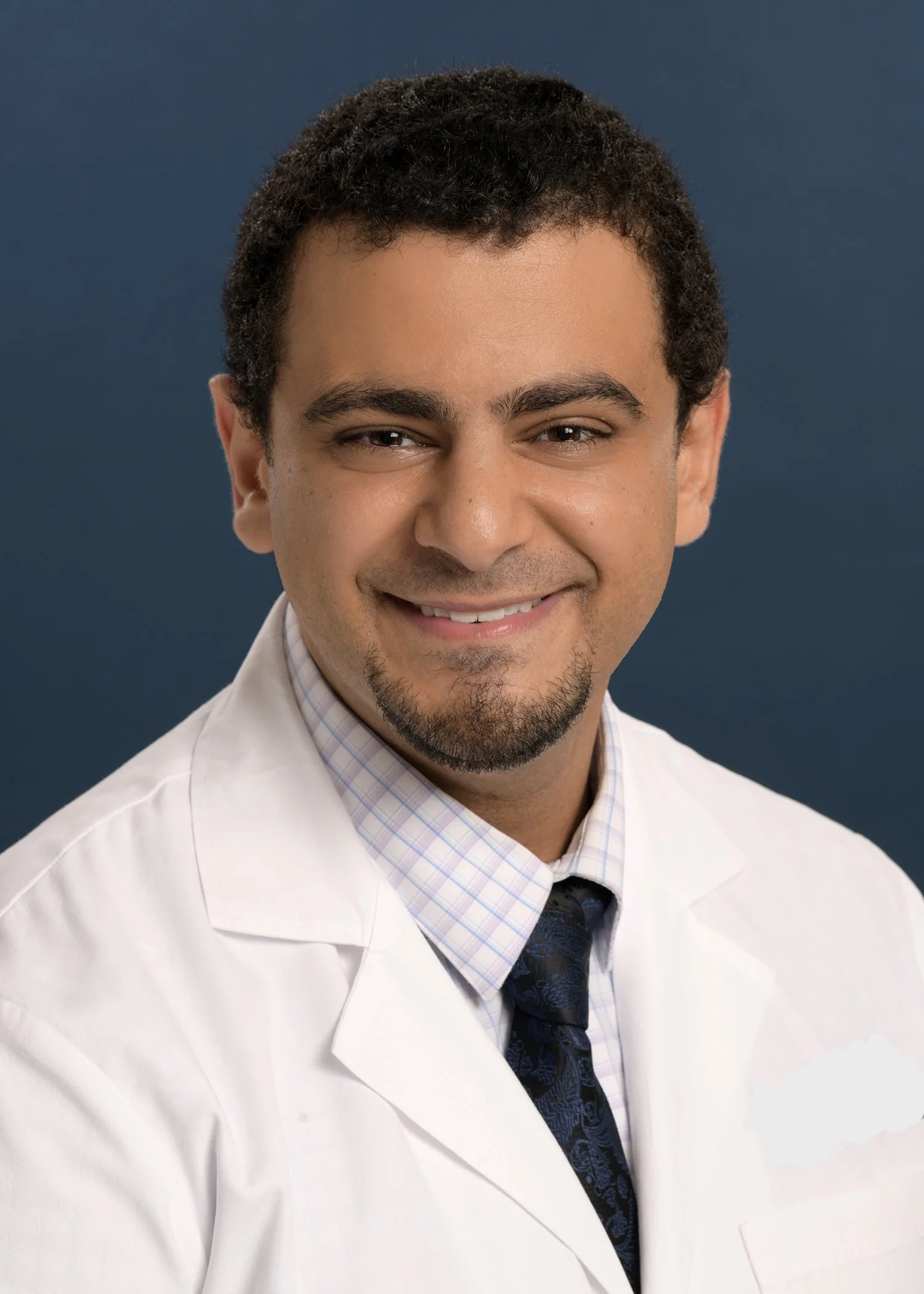 Professional portrait of a smiling male doctor with dark curly hair and a goatee, wearing a white lab coat, checked shirt, and a dark patterned tie, against a dark blue background.