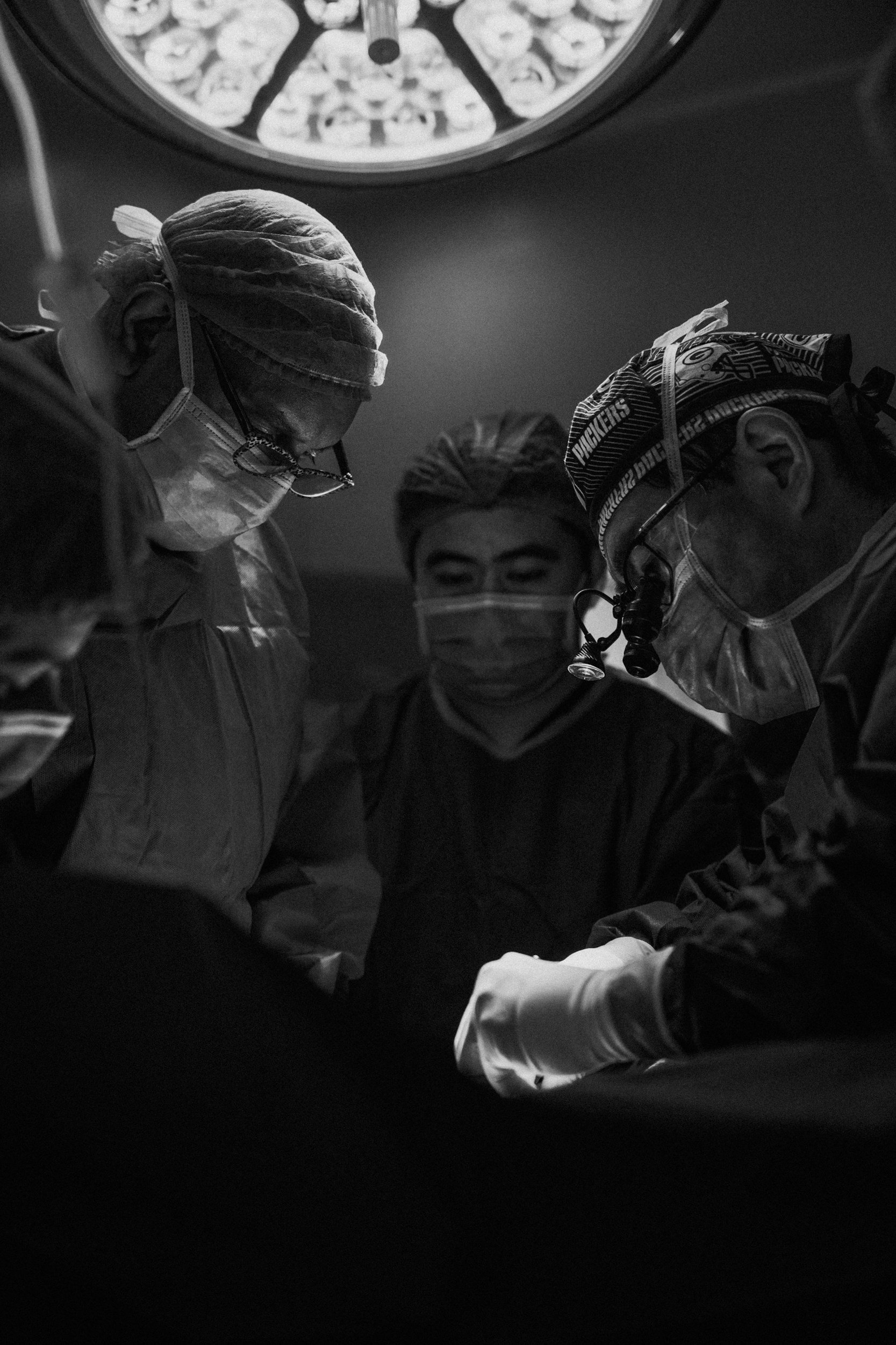 Medical team of four surgeons performing surgery, all wearing masks, surgical gowns, and head coverings, is seen from a low angle in an operating room, with a surgical light above.