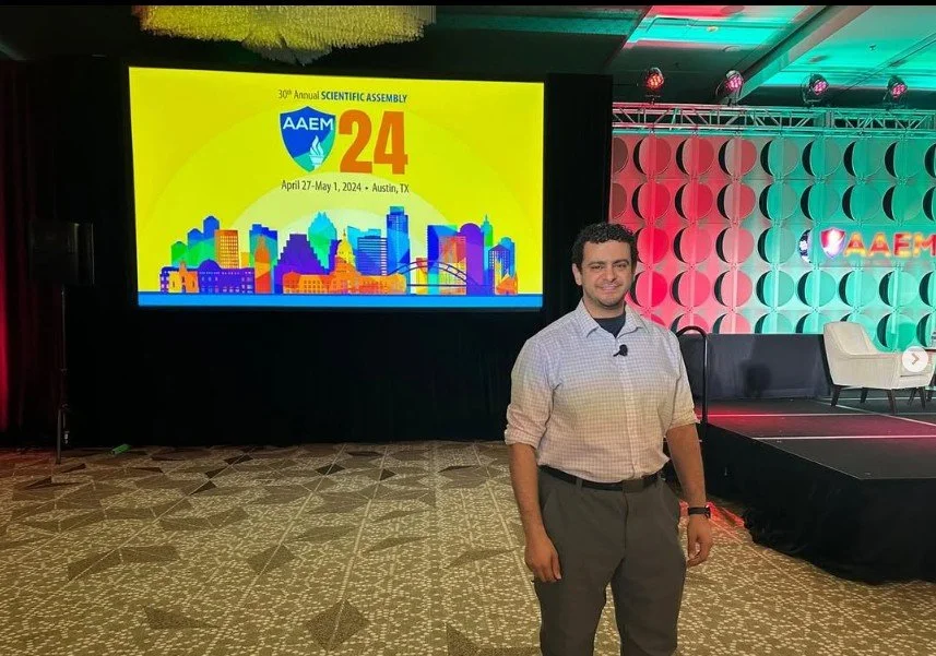 A man standing on a stage in front of a large screen displaying the AAEM 24th Annual Scientific Assembly logo, dates April 27-May 1, 2024, in Austin, Texas, with a colorful cityscape graphic. The stage is decorated with patterned walls and chairs.