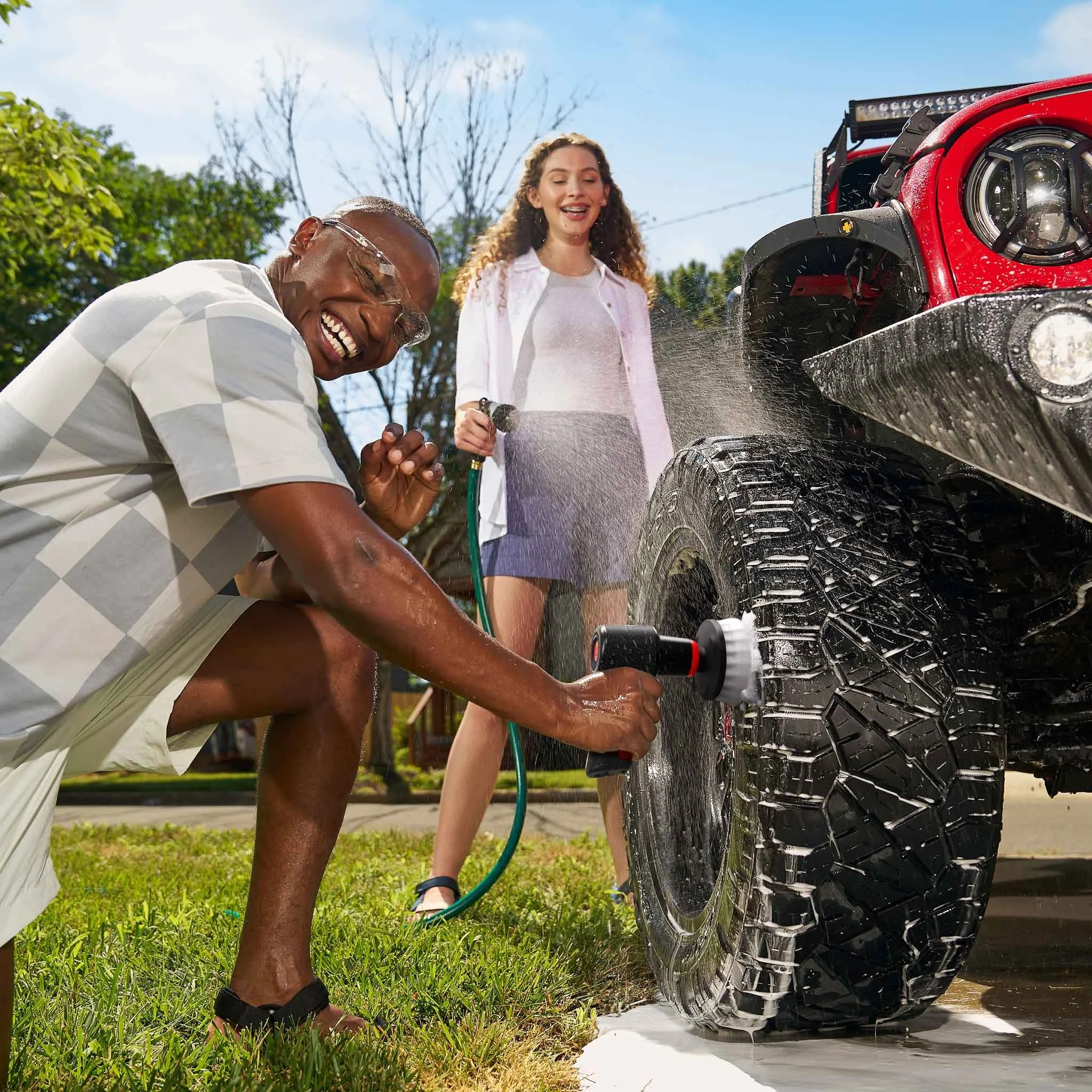 Dirt Devil handheld scrubber cleaning a jeep wheel. Woman spraying man with water hose.