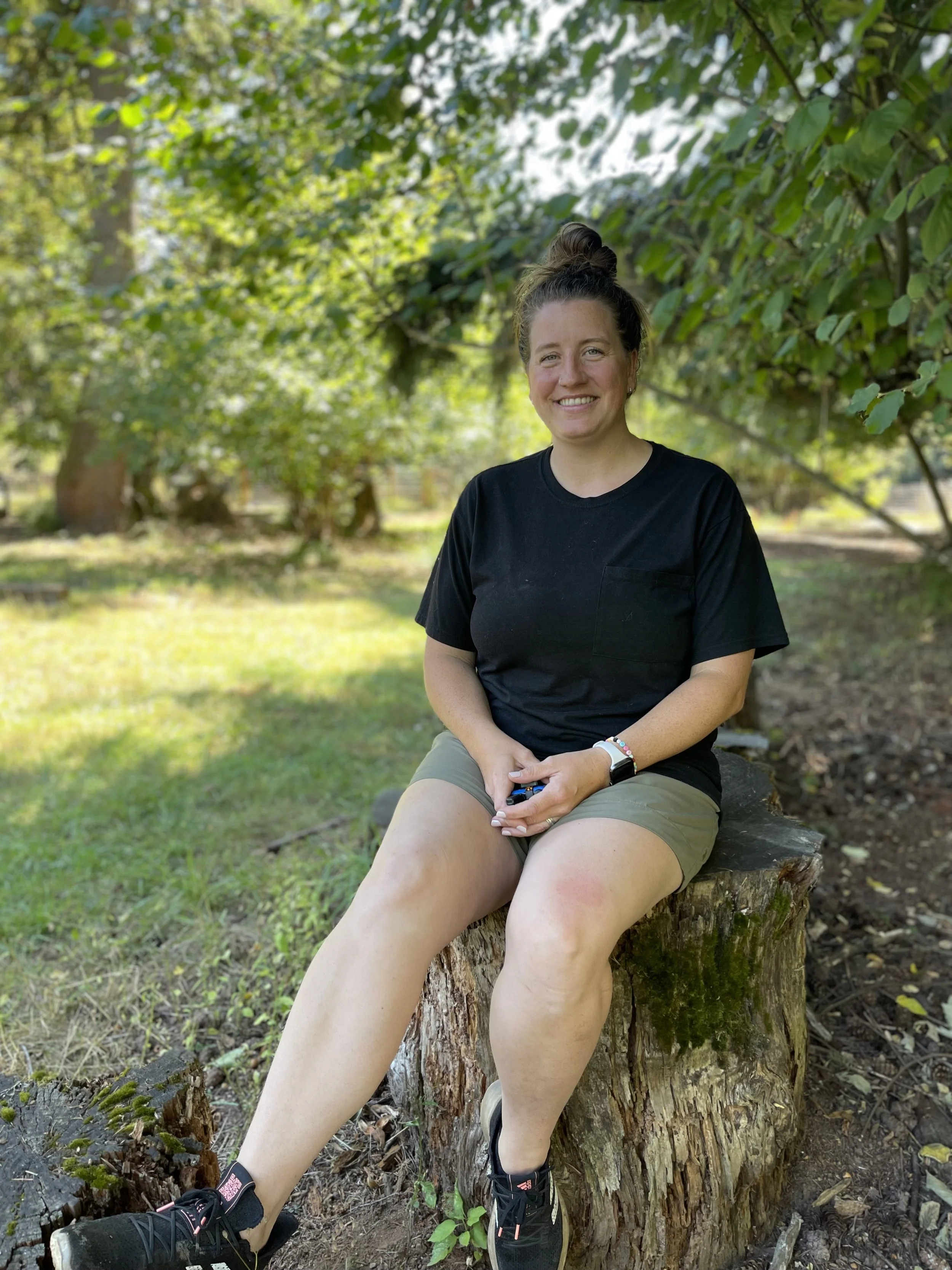 A young woman with brown hair tied up, sitting on a tree stump outdoors in a park, smiling, wearing a black T-shirt, khaki shorts, and black sneakers, surrounded by green trees and grass.