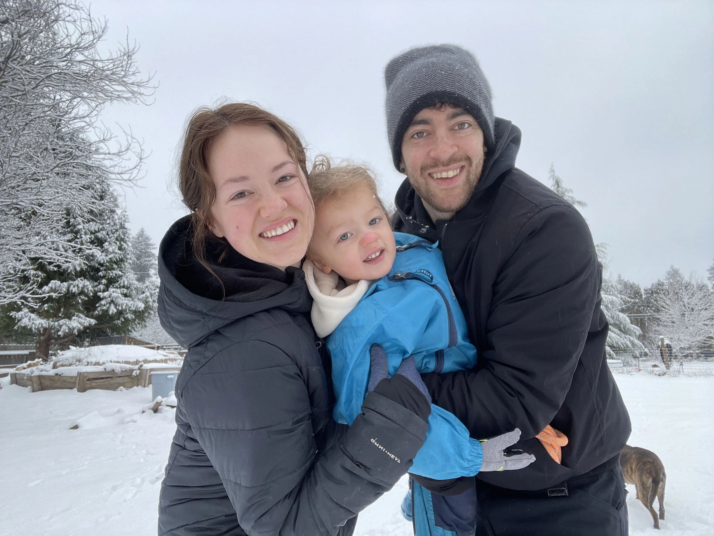 A smiling family of three, a woman, a man, and a young child, enjoying a snowy outdoor scene with snow-covered trees in the background.
