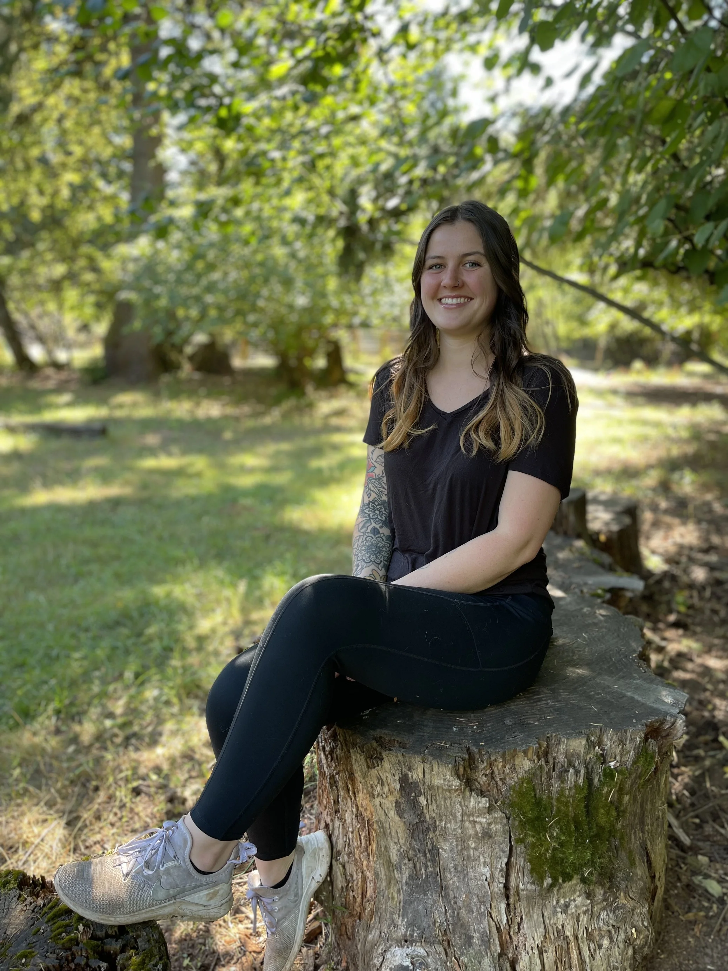 A young woman with long wavy hair, wearing a black t-shirt, black leggings, and beige sneakers, sitting on a large tree stump in a sunny park with green trees and grass in the background, smiling at the camera.