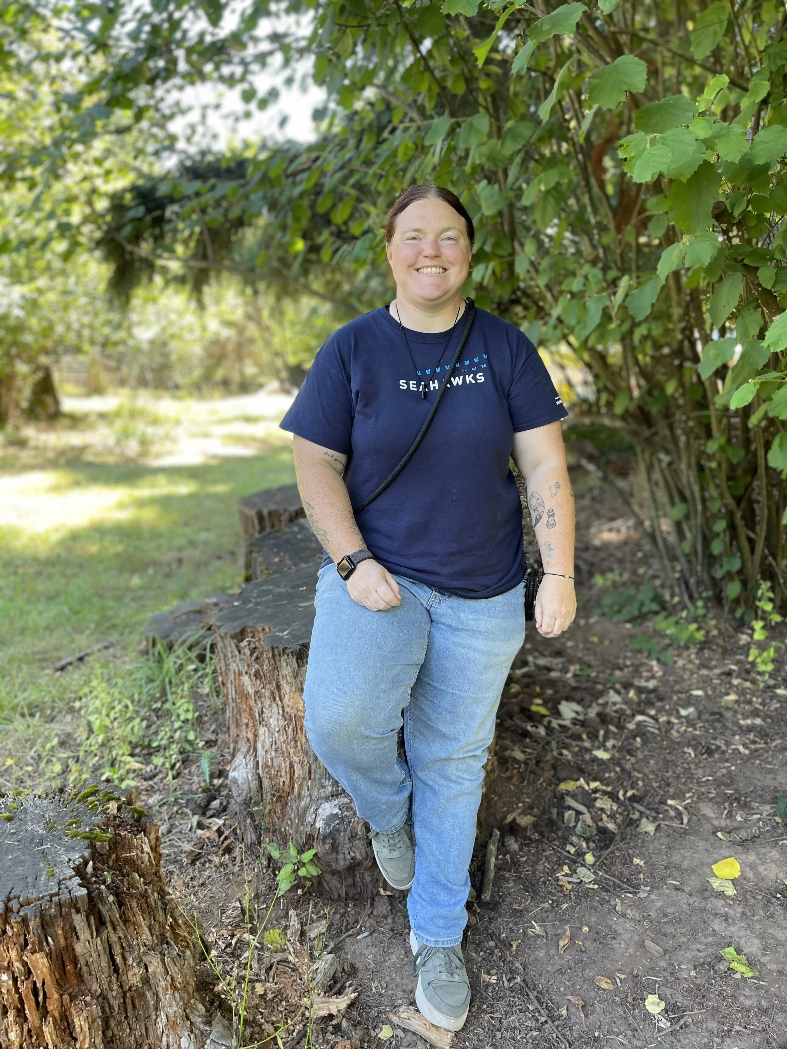 A woman standing outdoors on a trail surrounded by green foliage, leaning against a tree stump, smiling at the camera, wearing a navy blue t-shirt, blue jeans, and gray sneakers.