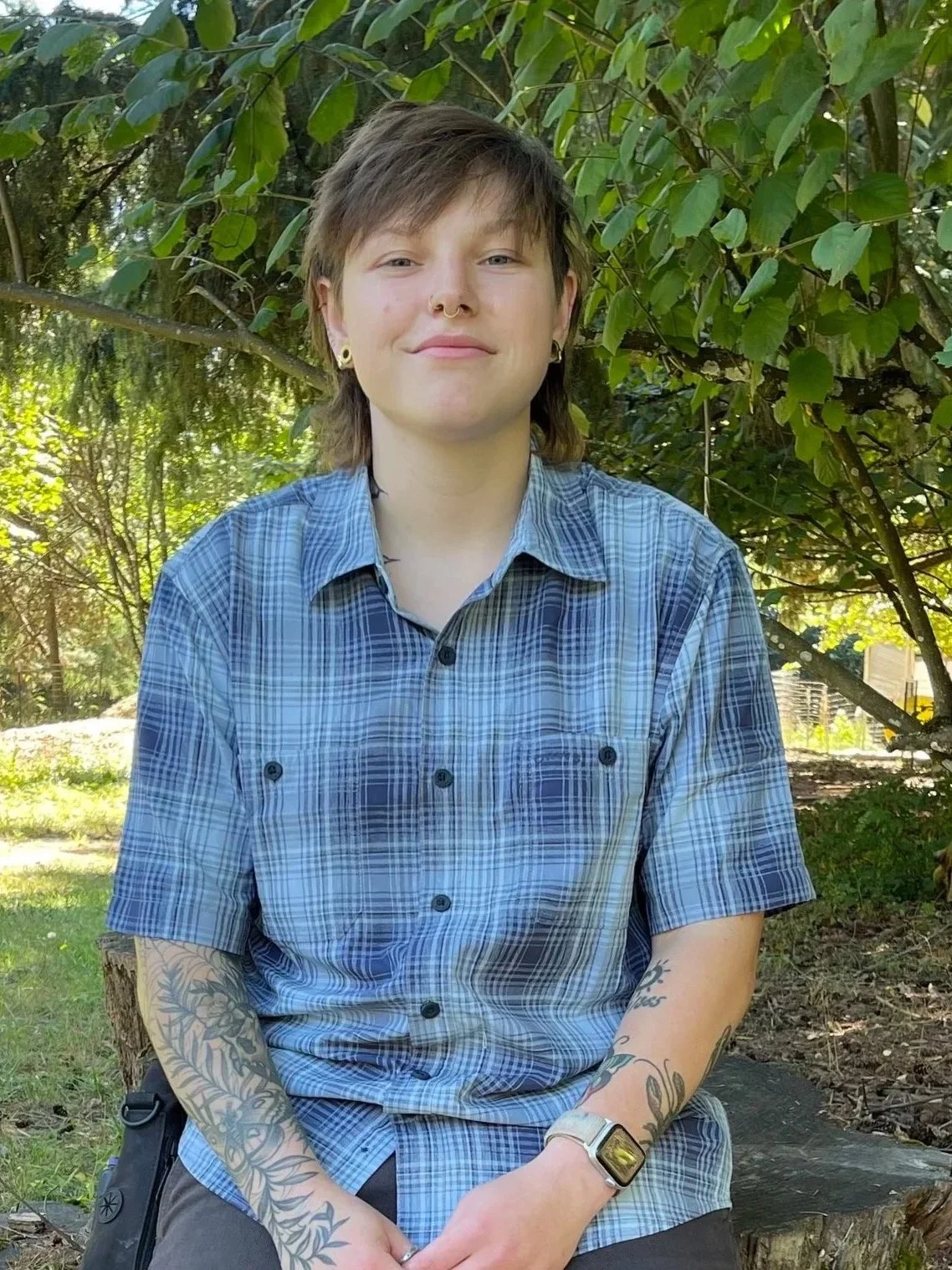 A young person with short brown hair, tattoos on arms and legs, wearing a blue plaid shirt, shorts, and black boots, sitting on a tree stump in a lush green outdoor setting.