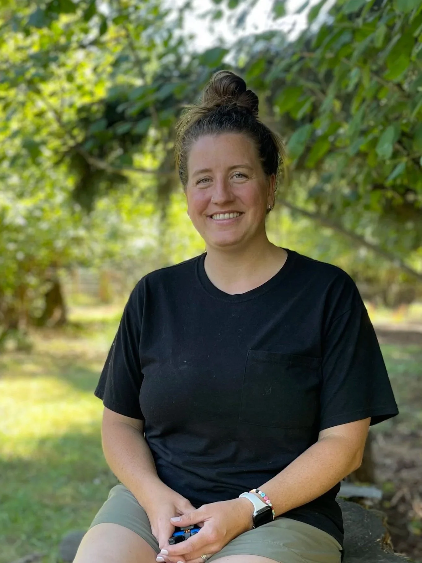 A young woman with brown hair tied up, sitting on a tree stump outdoors in a park, smiling, wearing a black T-shirt, khaki shorts, and black sneakers, surrounded by green trees and grass.