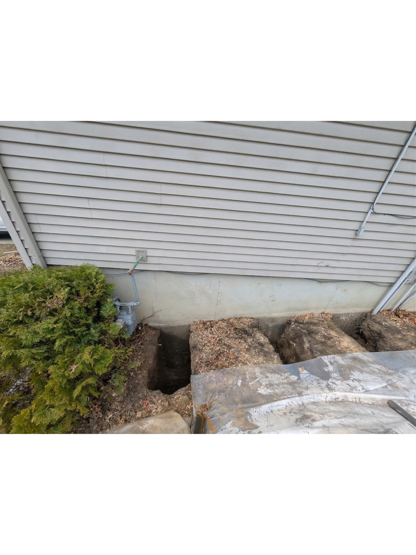 Outdoor area with a partially dug trench in front of a house with vinyl siding, a bush on the left, and dug-up soil and rocks.