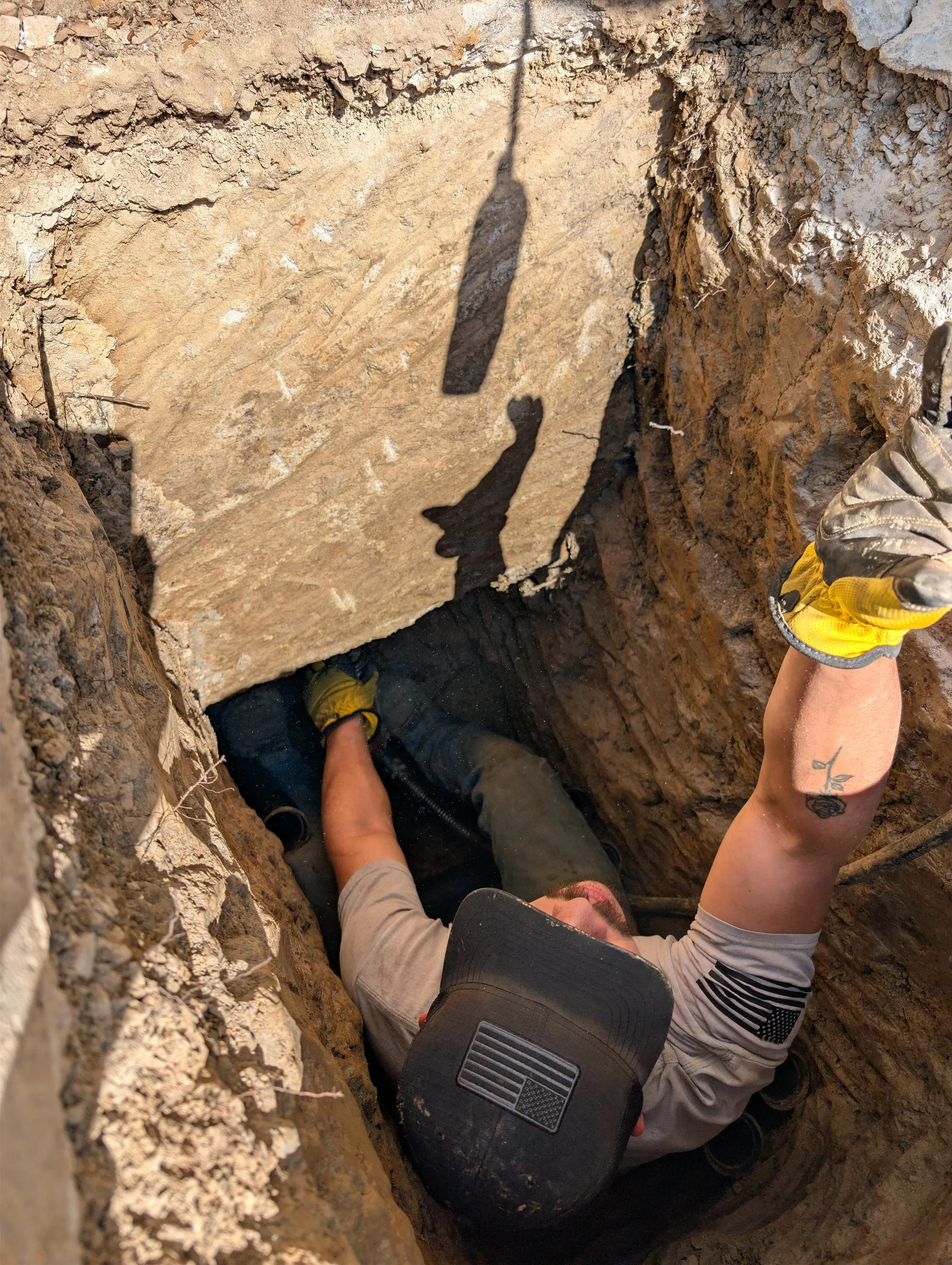 A person is crawling inside a narrow underground trench or hole, wearing gloves and a cap, with part of their body visible.