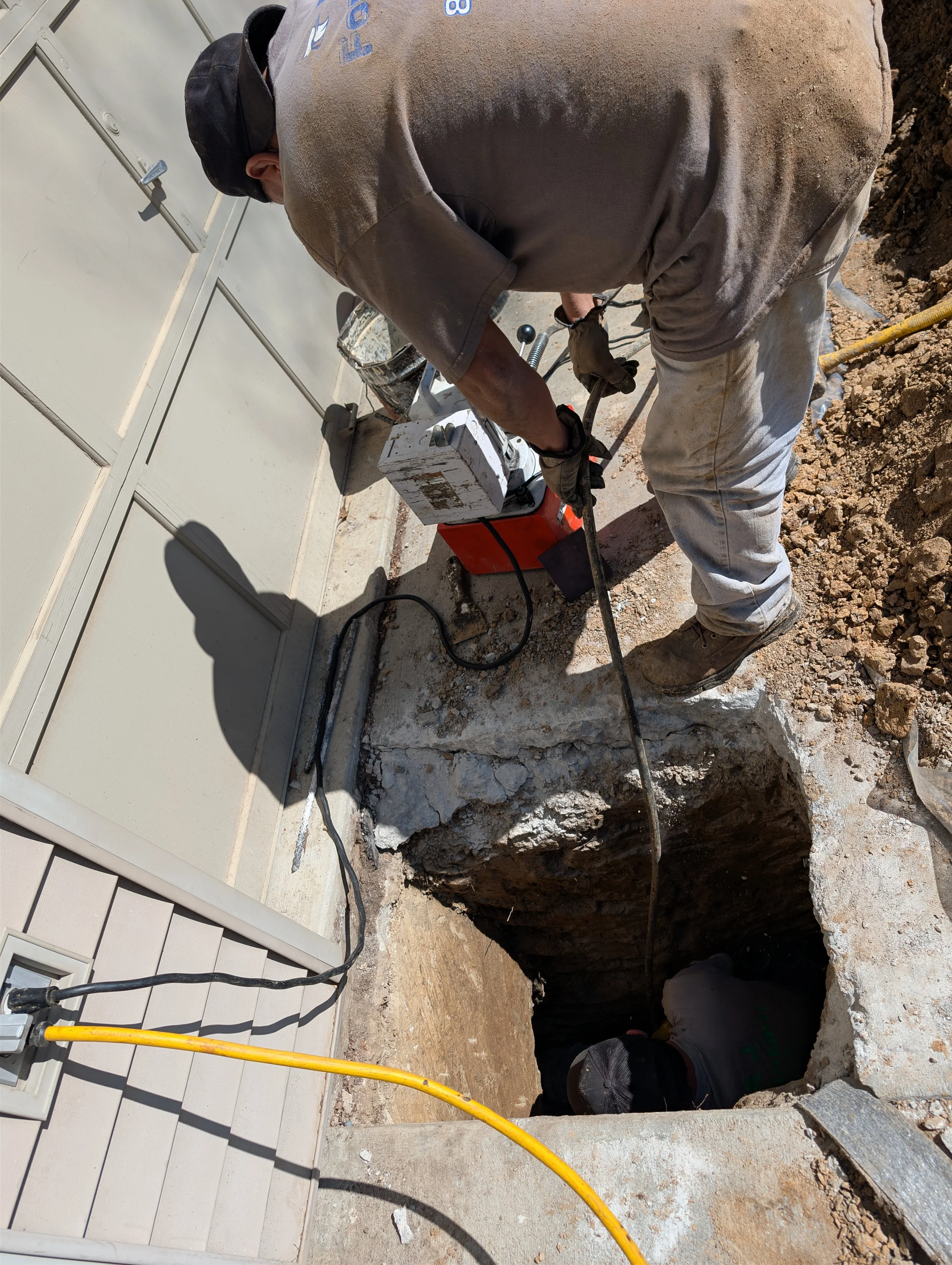 Construction worker operating a jackhammer in an excavated hole next to a house wall, with another worker visible inside the hole.