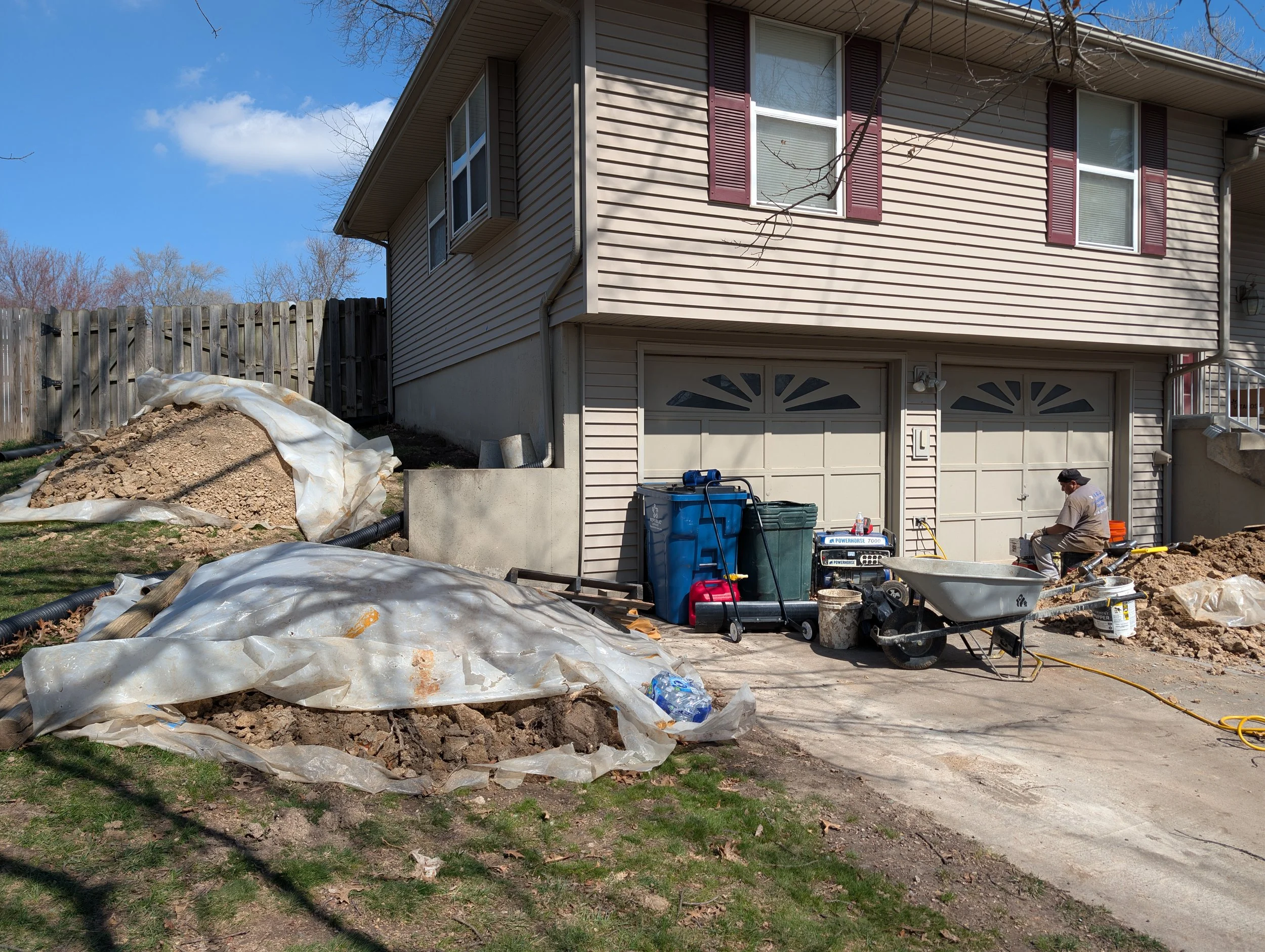 A construction worker working outside of a beige house with a double garage, surrounded by dirt and construction materials, including a wheelbarrow, plastic sheeting, and debris.